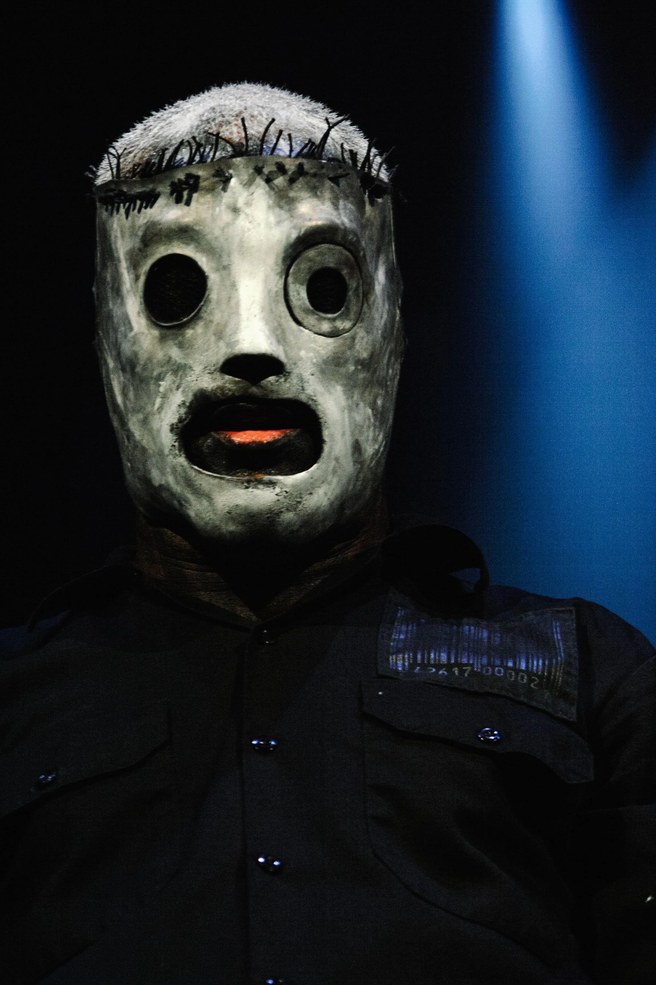 A person wearing a white, hollow mask with black eye holes and an open mouth. The mask has a crown made of small twigs or branches on top. The person is dressed in a dark shirt with a name tag and is illuminated by a blue spotlight from the side, set