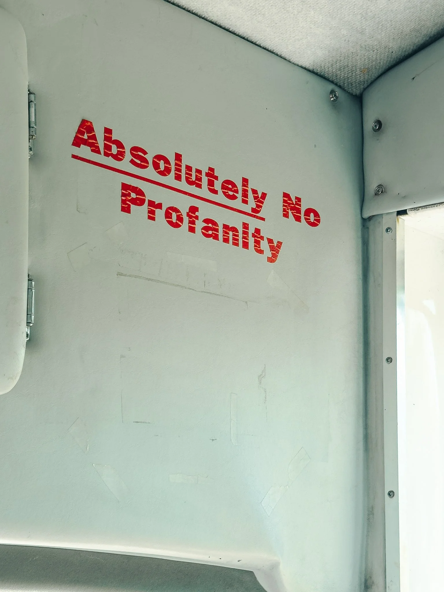 A white wall with a red painted sign that reads 'Absolutely No Profanity' with a line underneath. The wall has some scratches and a partial corner of a window or door frame on the right side.