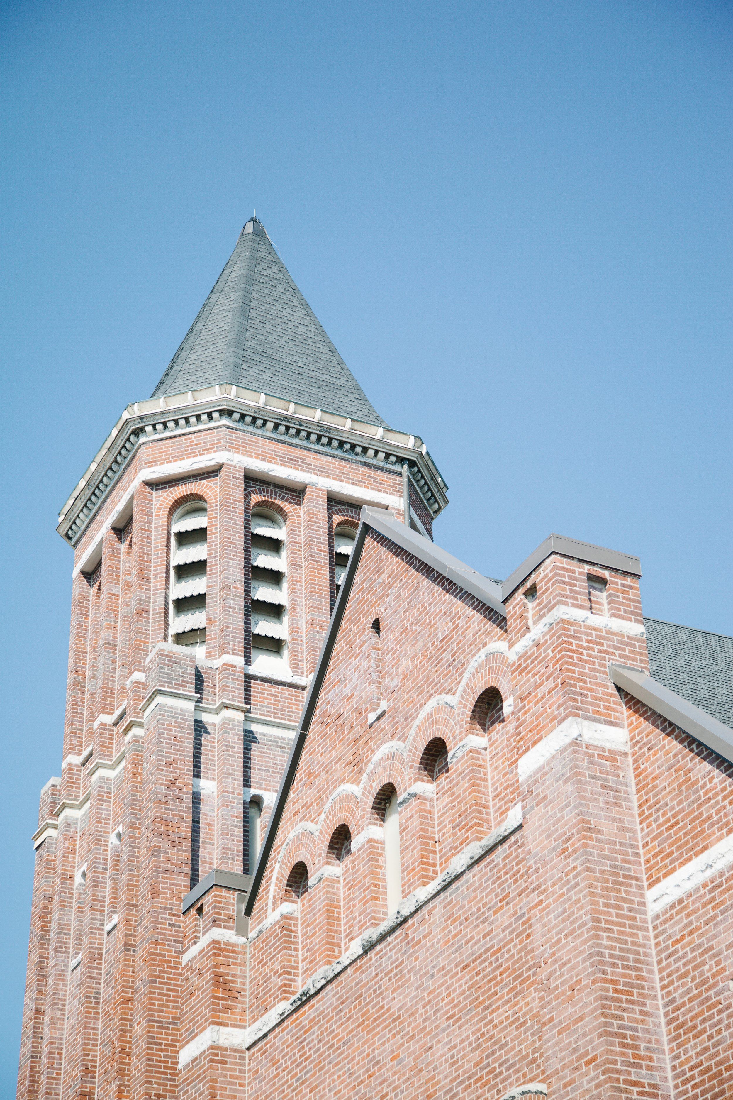 Upward view of a red brick church tower with arched windows and a pointed gray slate roof against a clear blue sky.