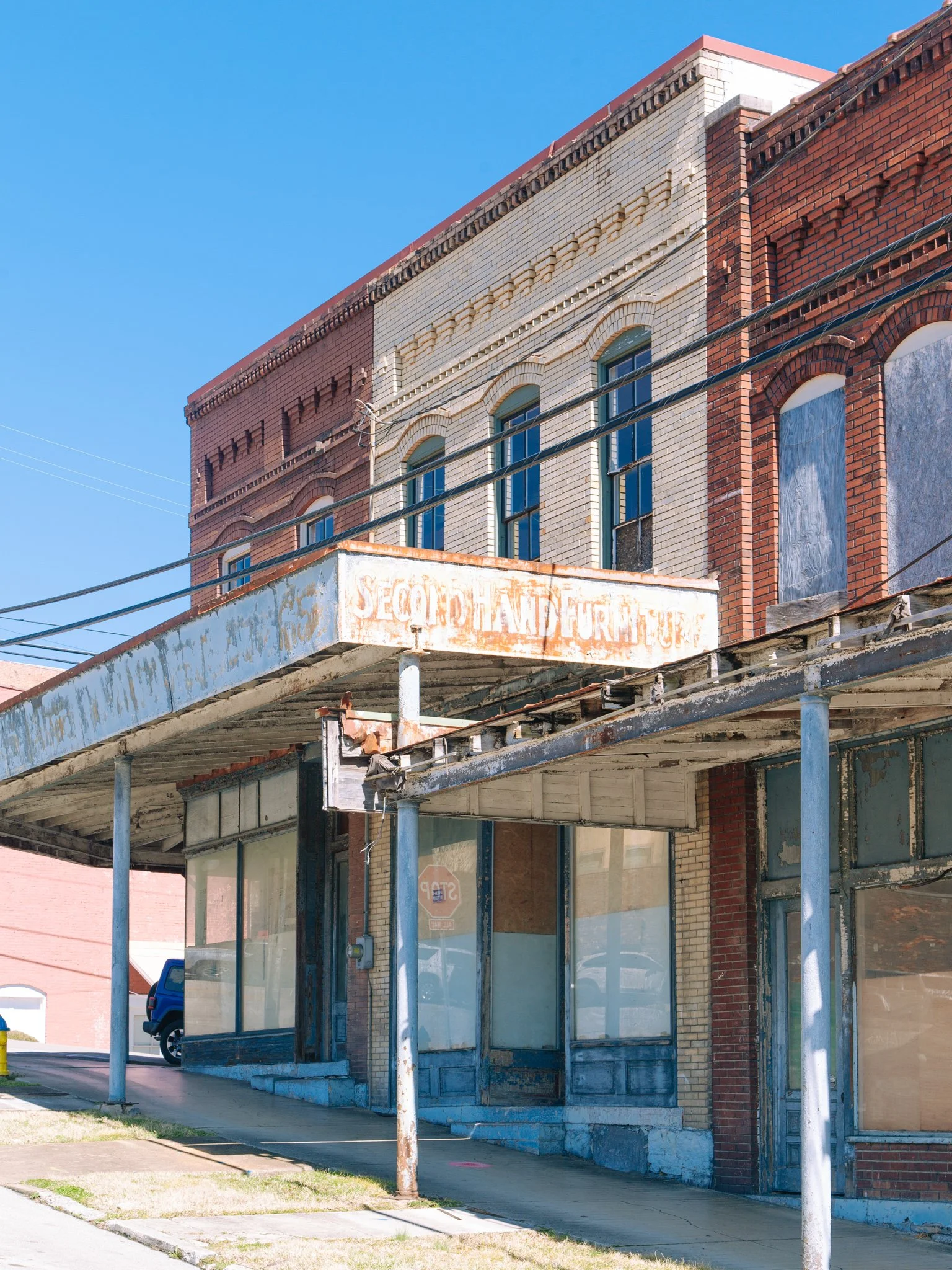 An old, abandoned storefront with boarded-up windows, a faded sign reading 'Secondhand Furniture,' a cracked sidewalk, and a nearby parked vehicle under a blue sky.