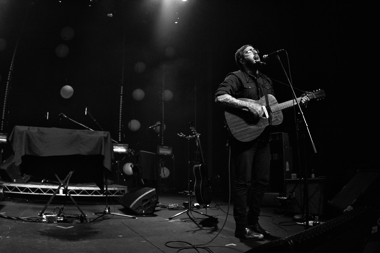 A man playing guitar and singing into a microphone on stage at a concert, with music equipment and lighting in the background.