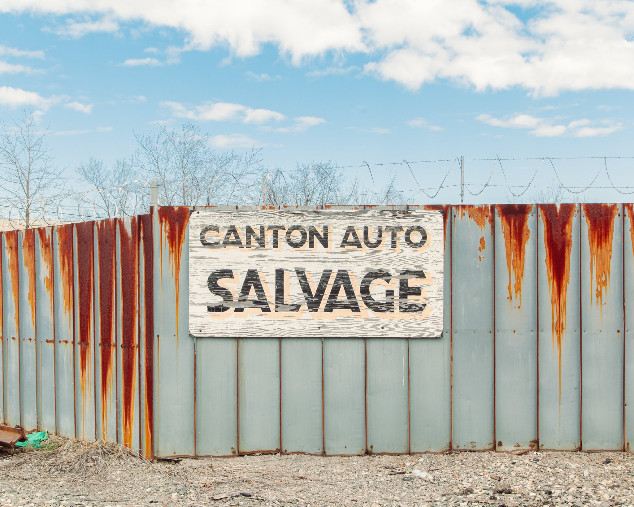 Rusty metal fence with a worn wood sign in front of leafless trees under a partly cloudy sky.