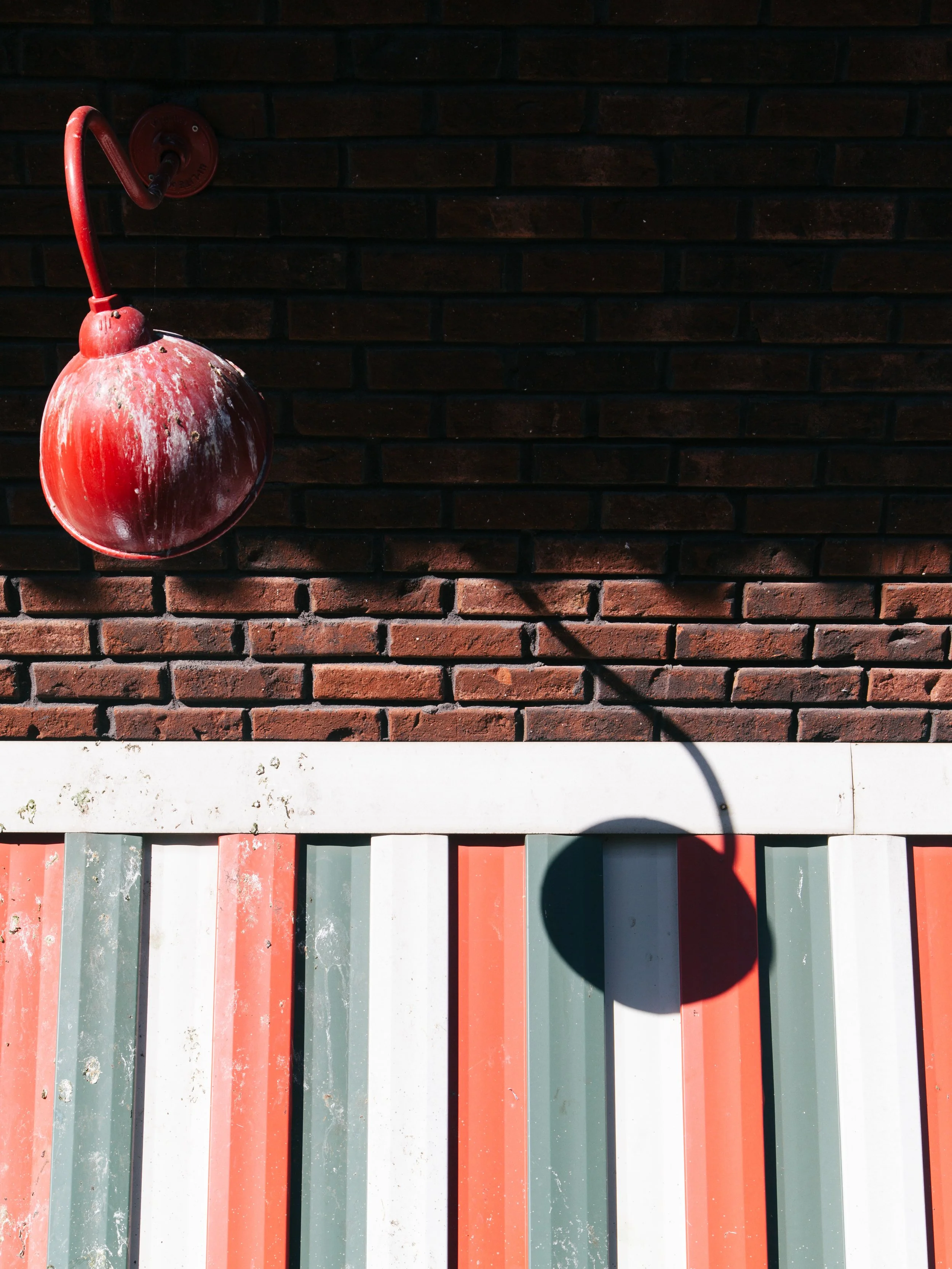 A red, weathered fire hydrant attached to a brick wall, casting a shadow on a colorful fence below.