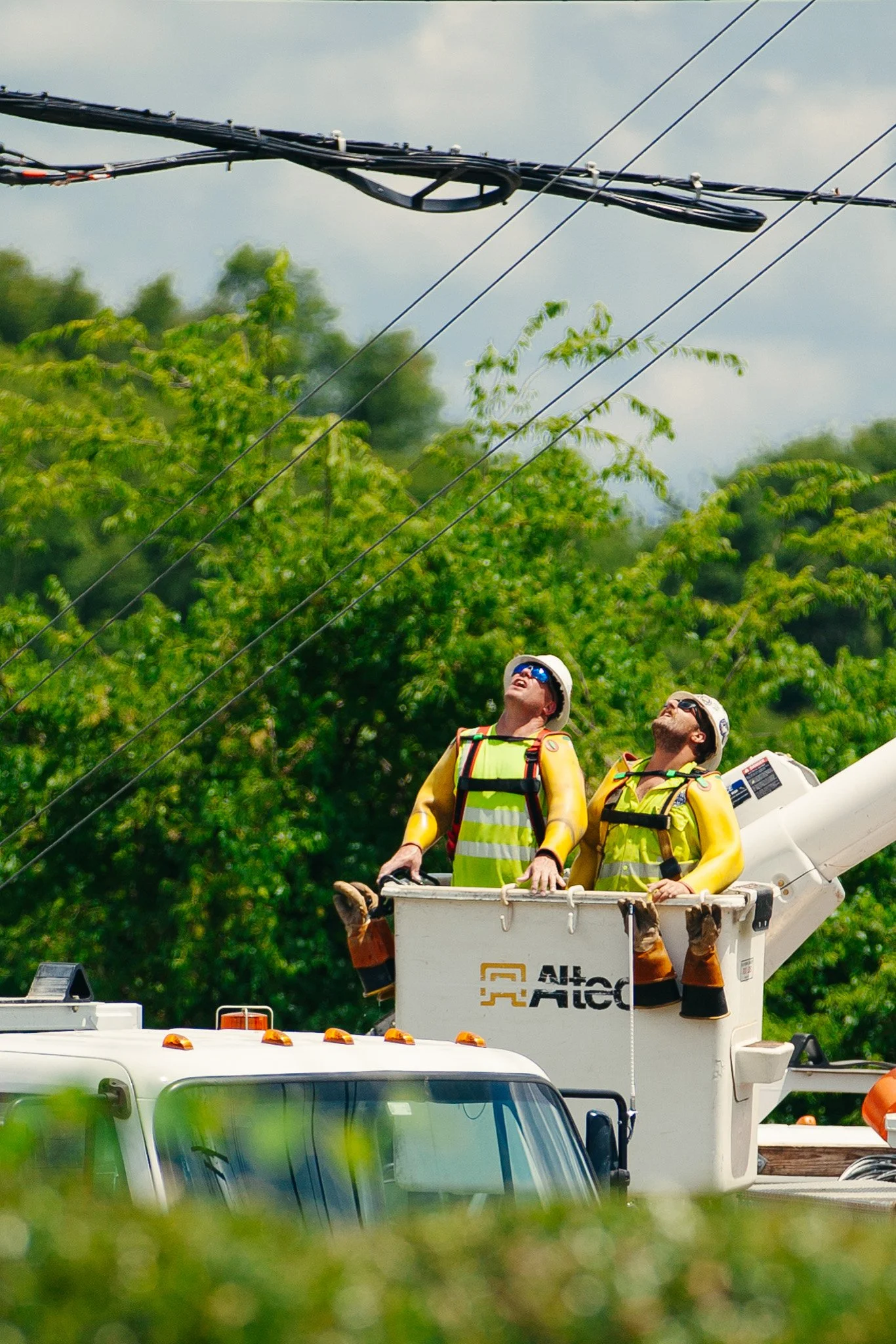 Two utility workers in yellow safety vests and helmets stand in an elevated bucket on a truck, working on electrical power lines among green trees under a cloudy sky.