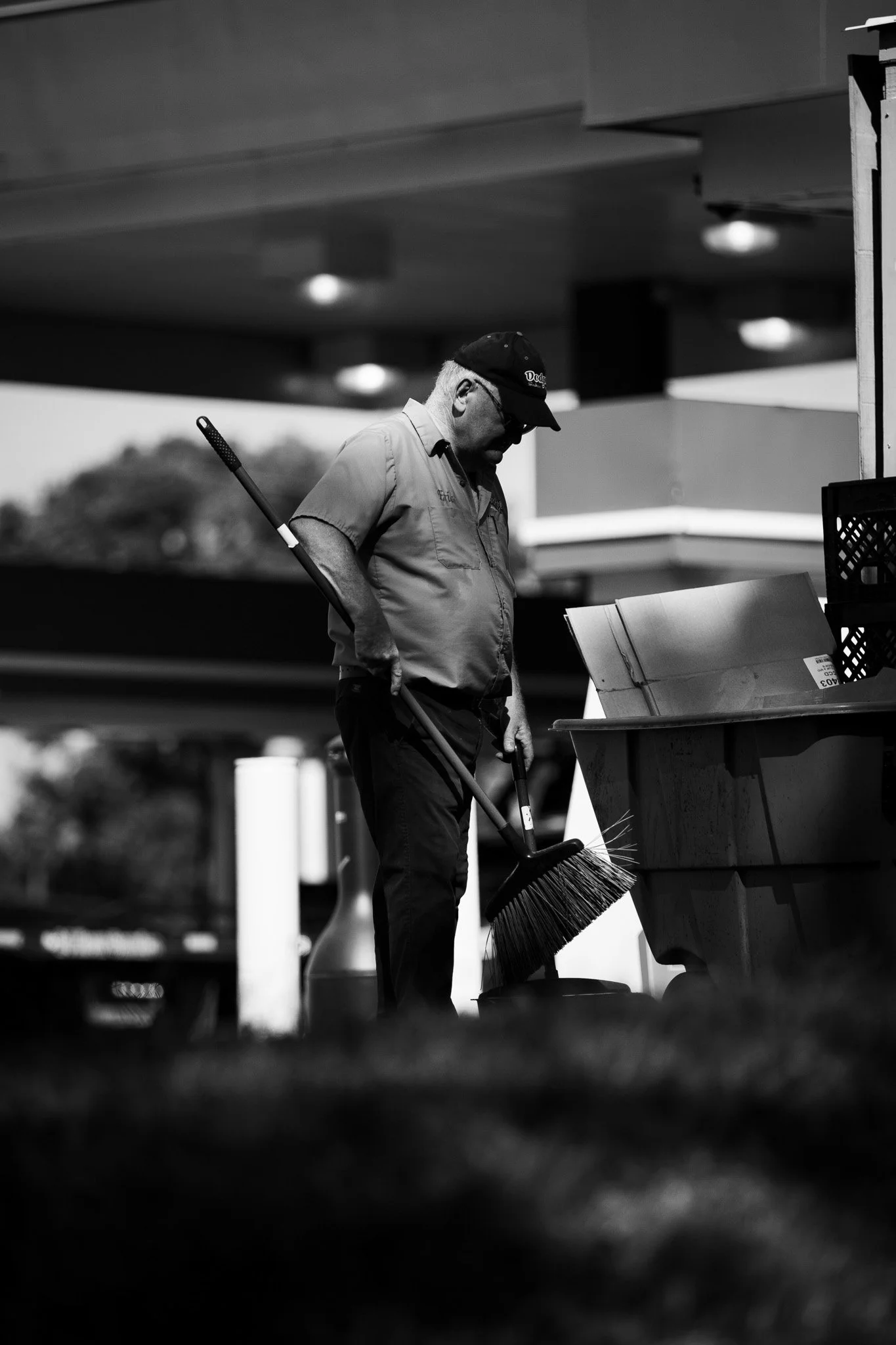 A man wearing a uniform, cap, and glasses stands in front of a trash bin with a broom, looking down, possibly cleaning outside a building, in black and white.