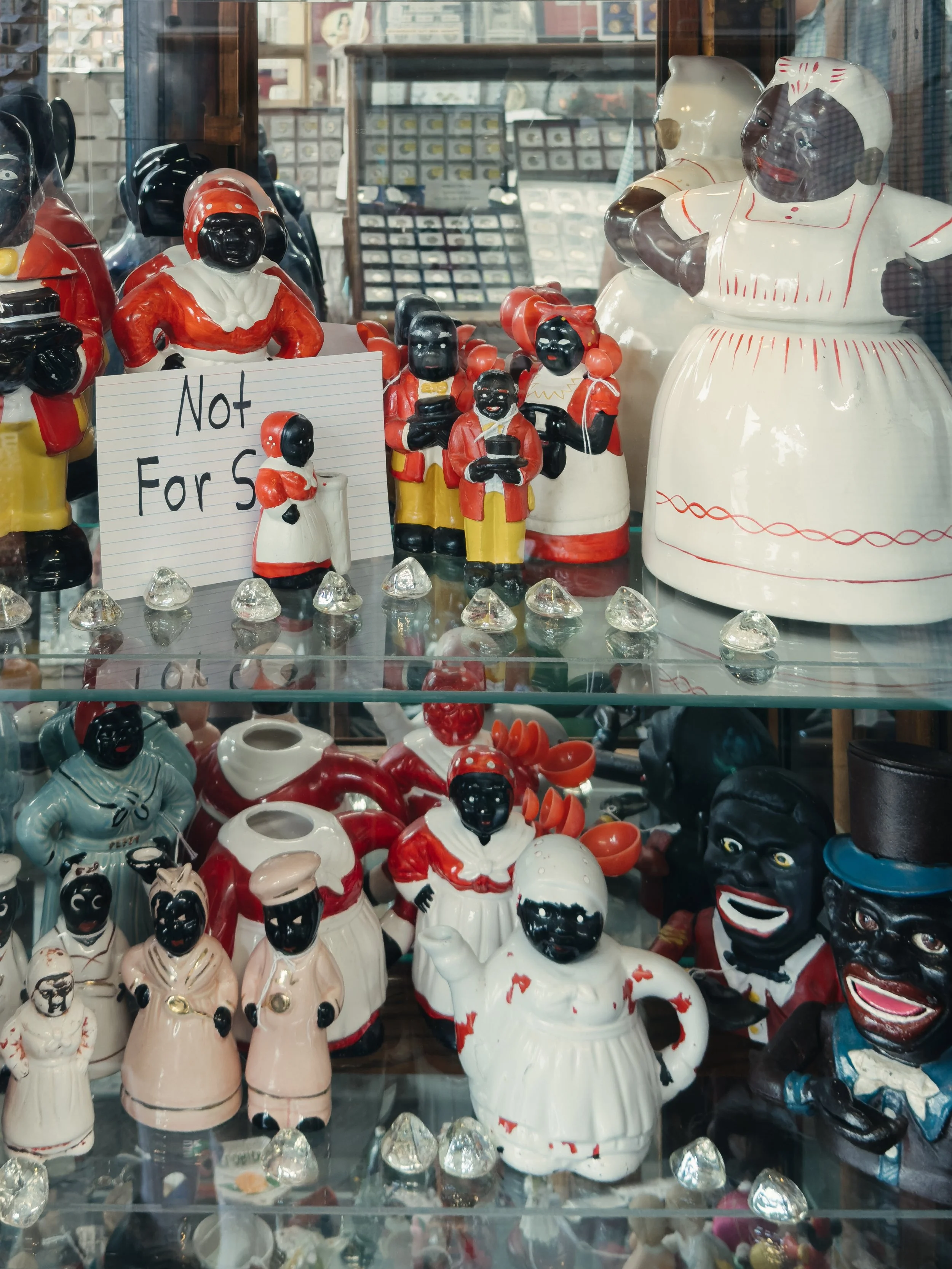A display of vintage ceramic figurines of Black characters dressed in various costumes, some in traditional clothing and others in uniforms, inside a glass case at a store. There is a note on the display that says 'Not For Sale'.