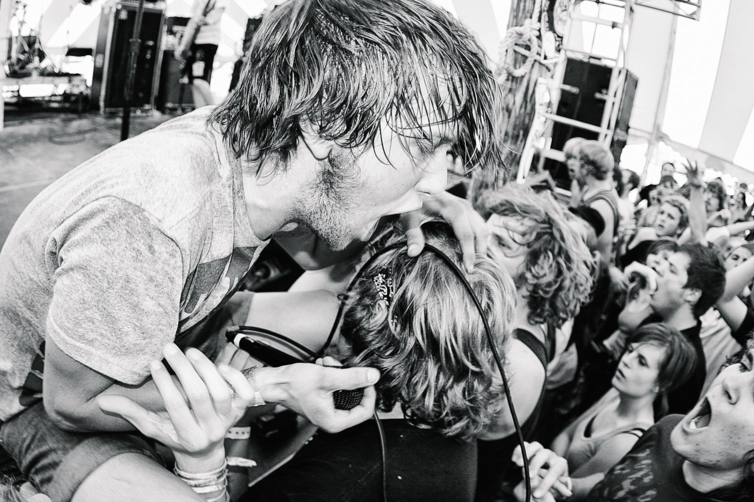 A man with long hair and a beard leaning over a woman at a concert, with his hand on her head, while holding a microphone. The crowd around them is excited, with some people singing and raising their hands.
