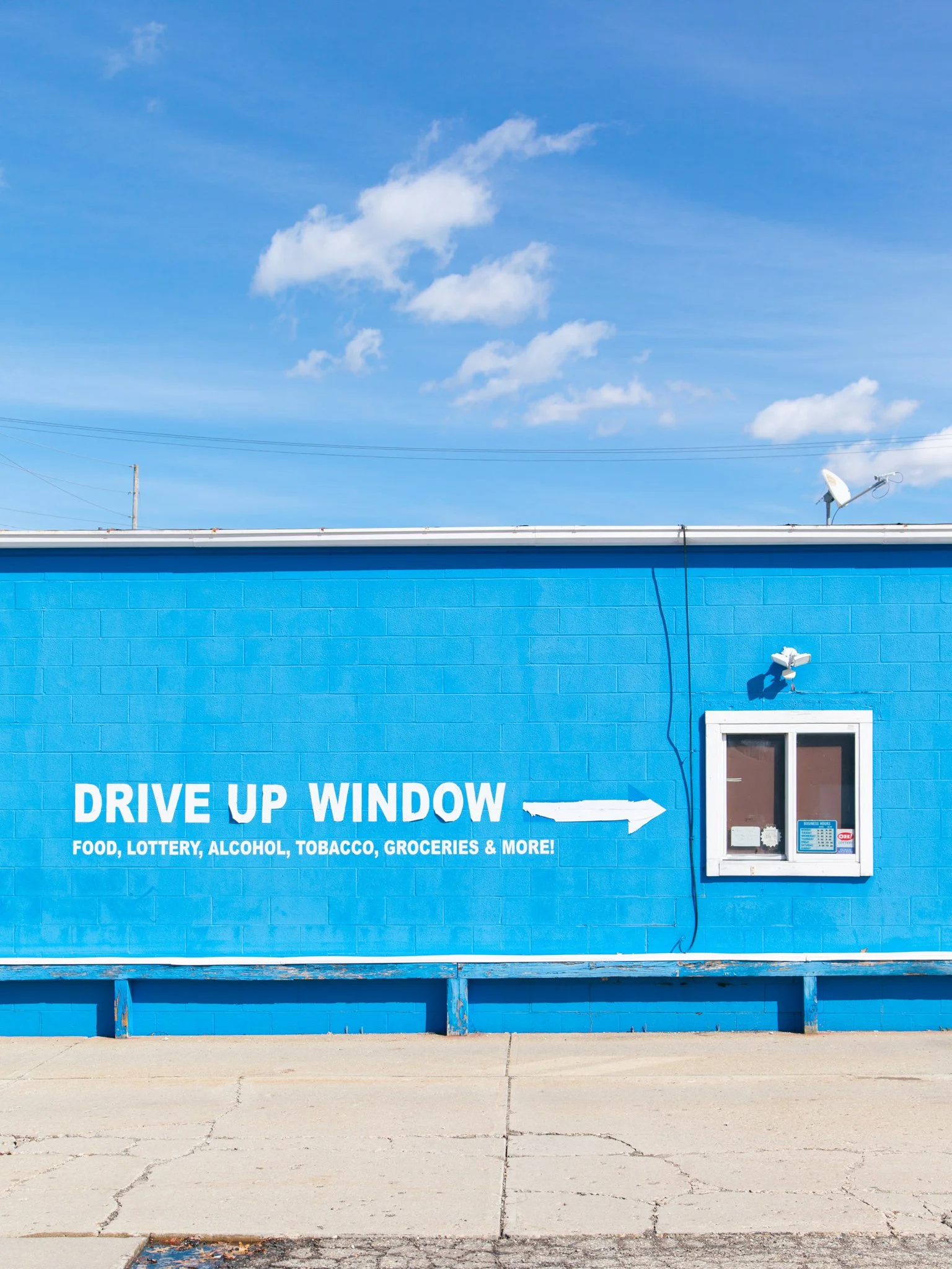 Blue building with a white window and a sign indicating drive-up window for food, lottery, alcohol, tobacco, groceries, and more, with a bright blue sky and a few clouds in the background.