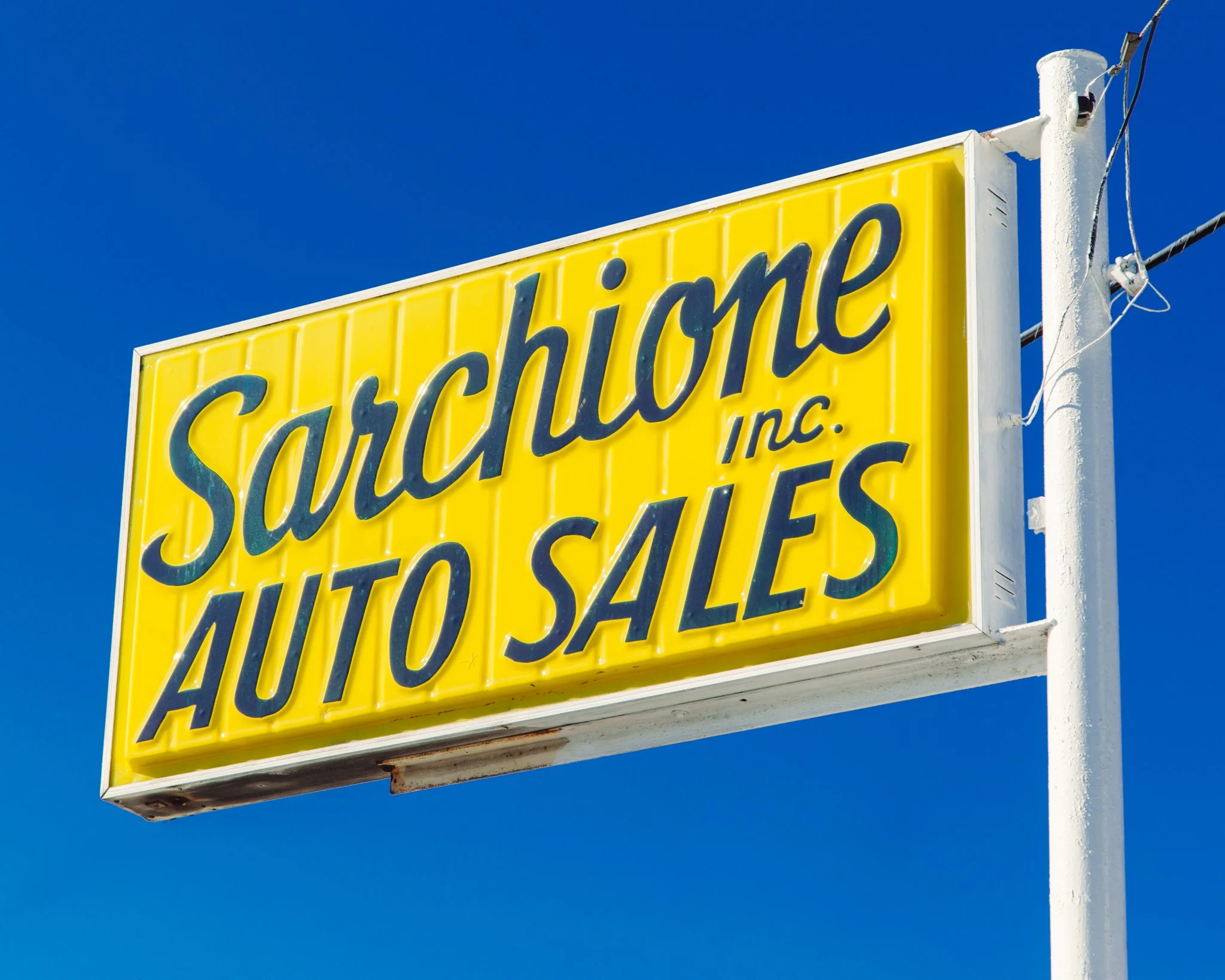 Close-up of a bright yellow sign with blue text that reads 'Salvation Inc.', mounted on a white pole against a clear blue sky.