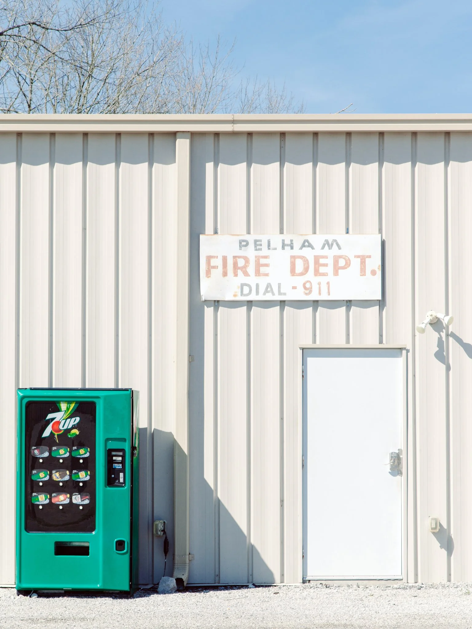 A white metal building with a sign that reads 'Pelham Fire Dept. Dial 911.' Next to it is a green 7UP vending machine. The sky is clear and there are leafless trees in the background.