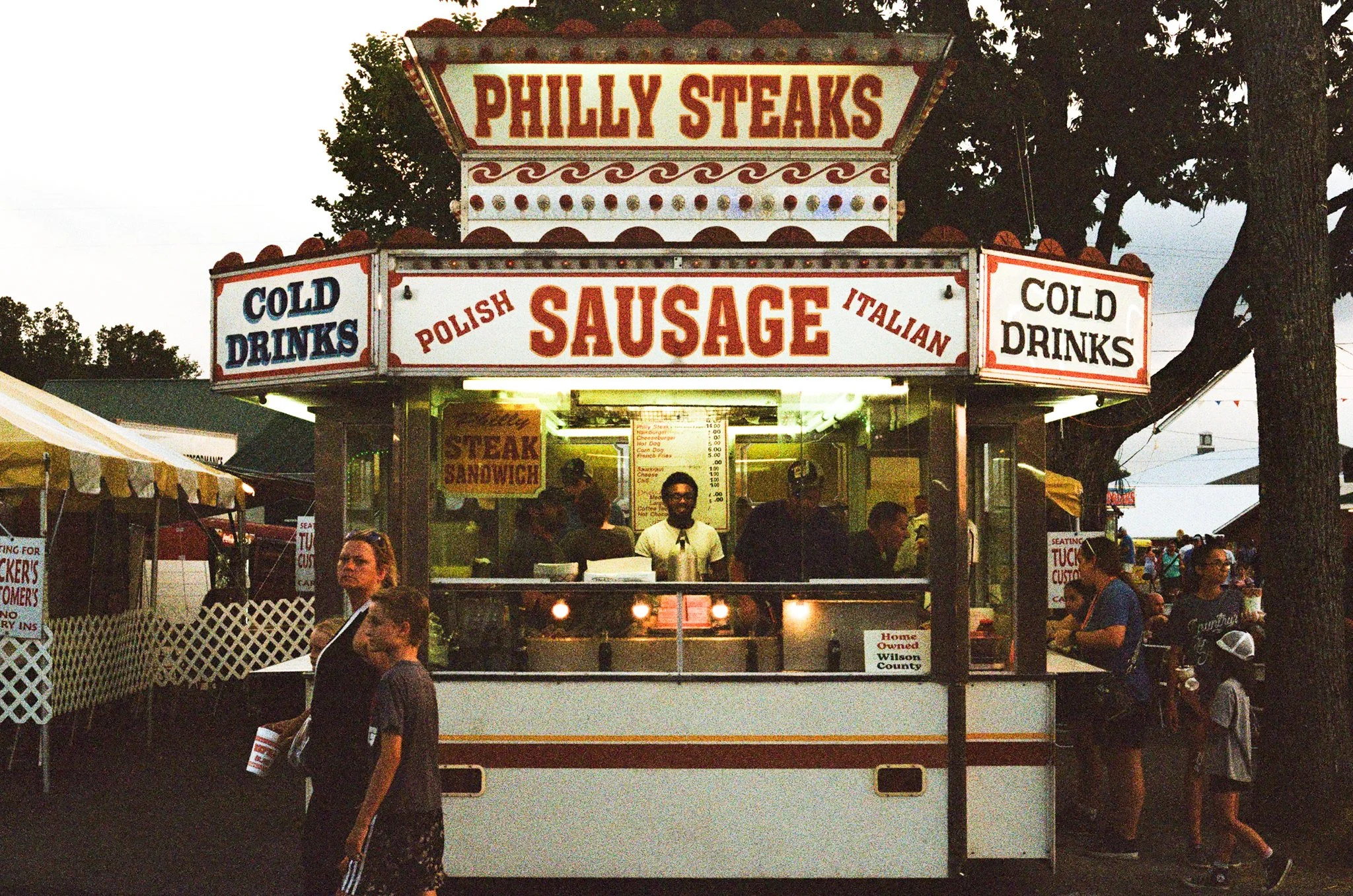 Food stand at a fair with signs advertising Philly steaks, Polish sausage, Italian sausage, and cold drinks. There are people walking and standing around, and trees are visible in the background during evening time.