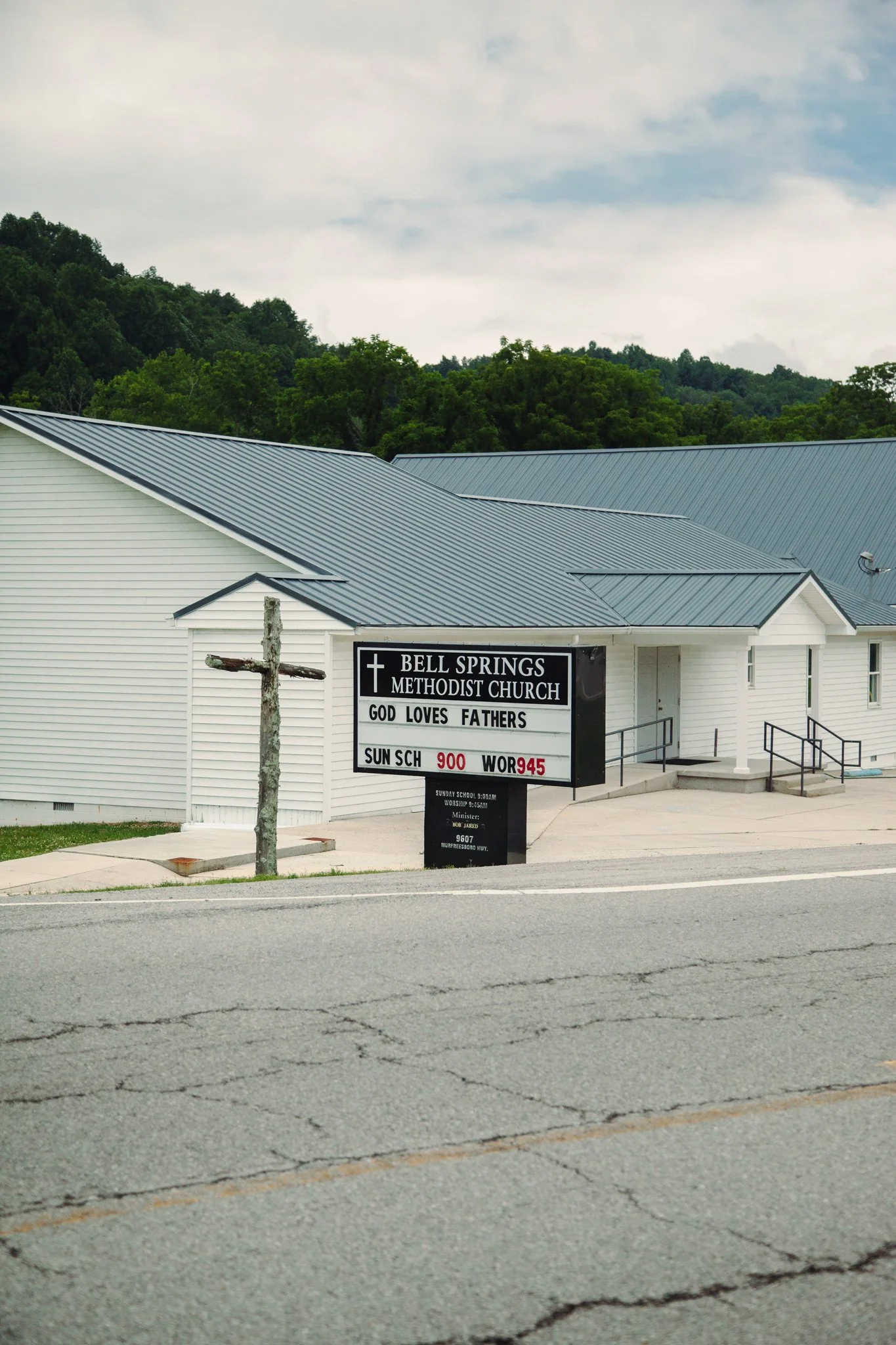 Front view of Bell Springs Methodist Church building with a sign in front indicating service times and religious messages, set against a background of green trees and cloudy sky.