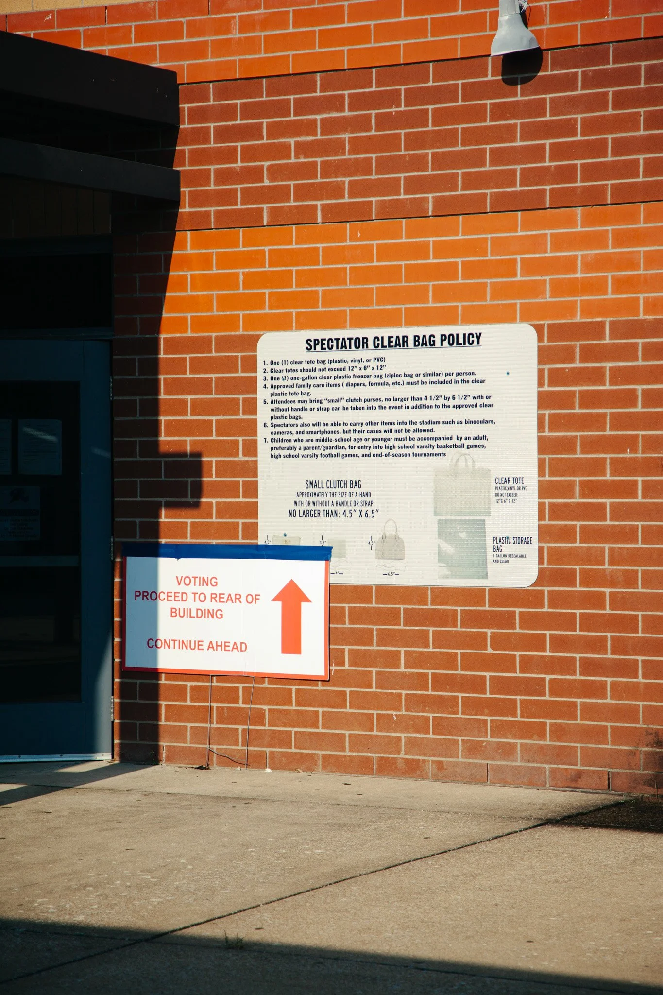 Sign on a brick wall outlining spectator clear bag policy, including acceptable bag types and size restrictions, and guidelines for bringing small clutch purses and other items into the stadium. Adjacent smaller sign with an arrow directs voters to p