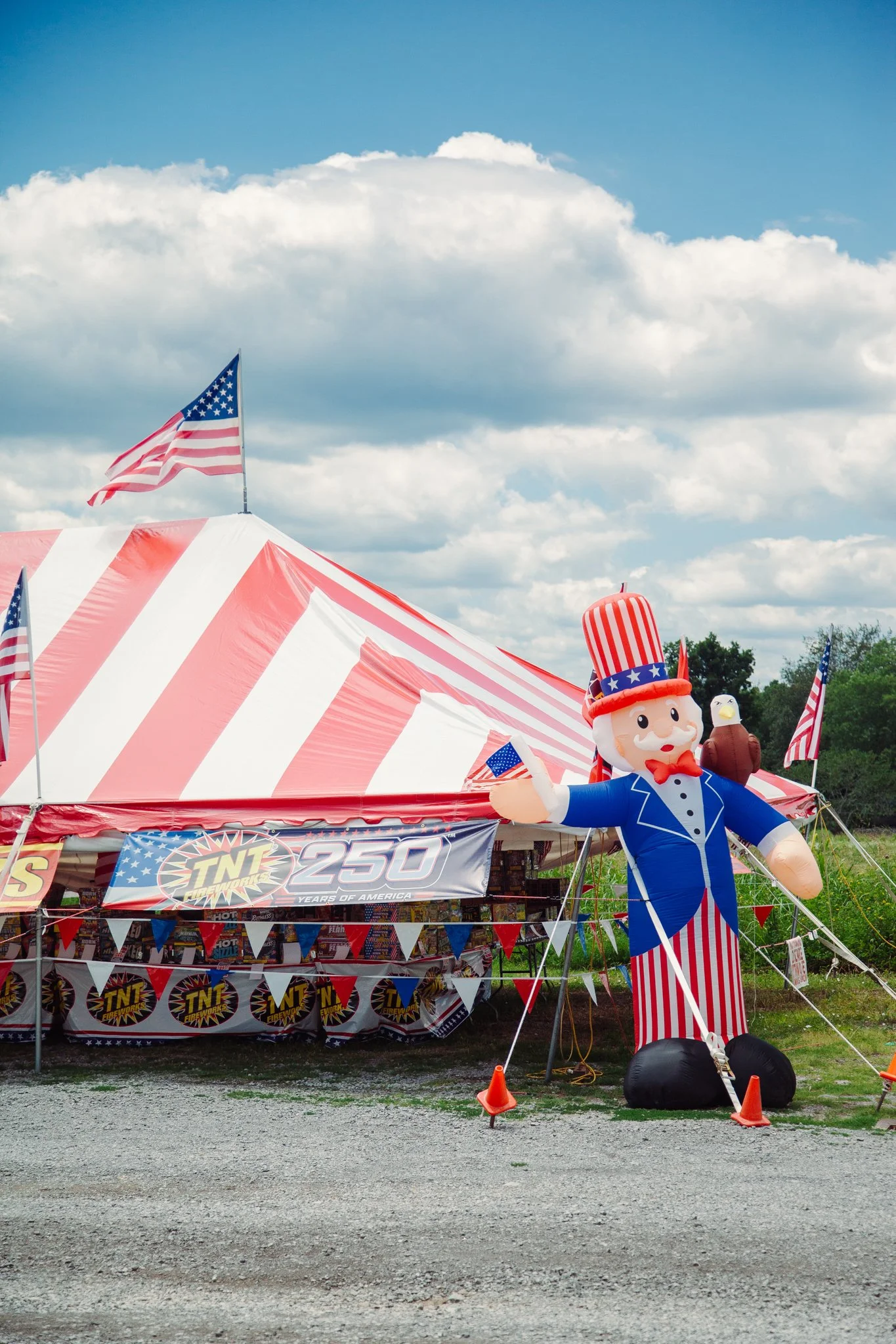 Red and white striped tent decorated with American flags, a large inflatable Uncle Sam figure, and banners celebrating 250 years of America, set outdoors on a sunny day with scattered clouds.