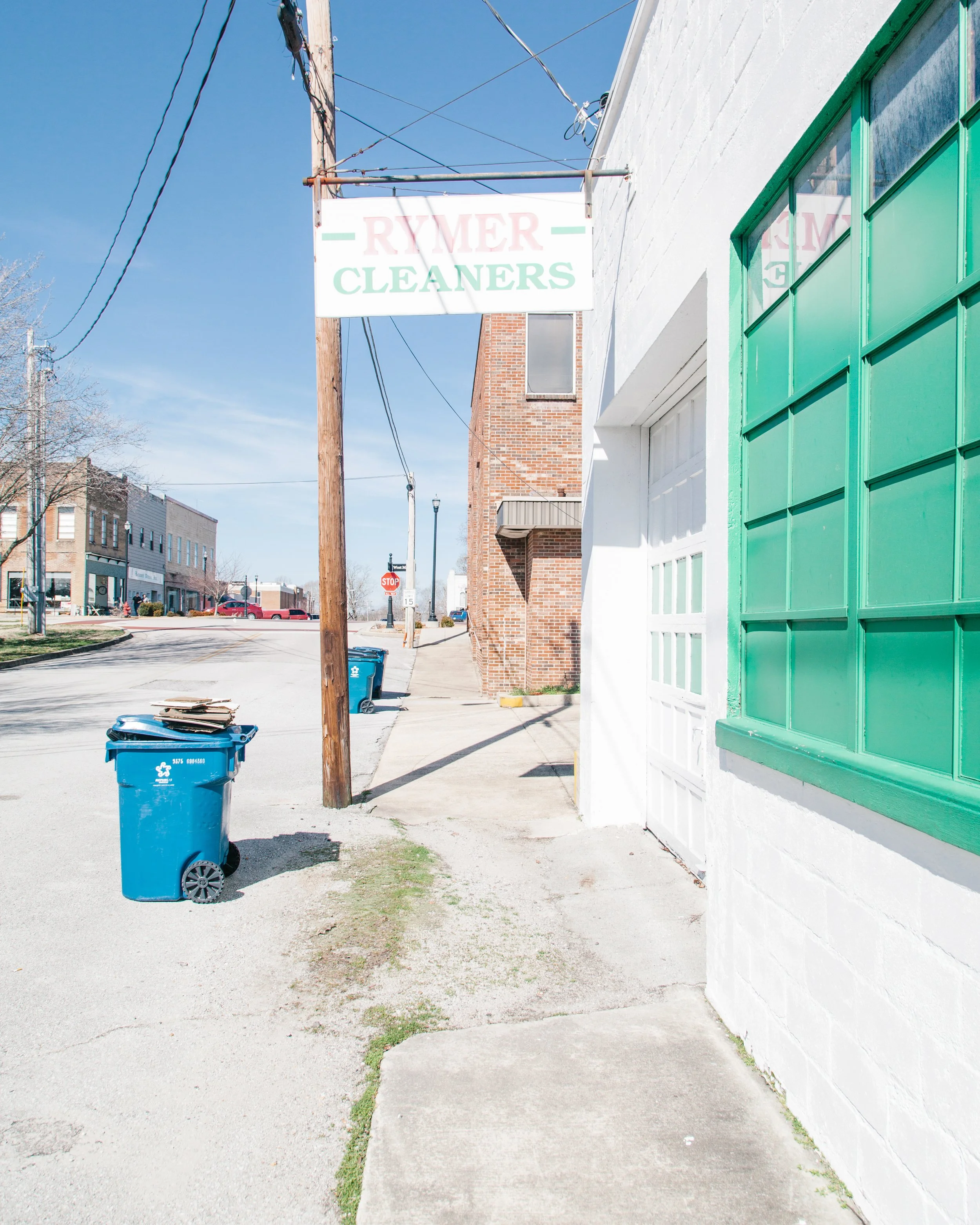 The image shows a sidewalk next to a white building with a large green window. There is a blue trash bin with some cardboard on top, and a wooden utility pole with overhead wires. A street with parked cars and a stop sign is visible in the background