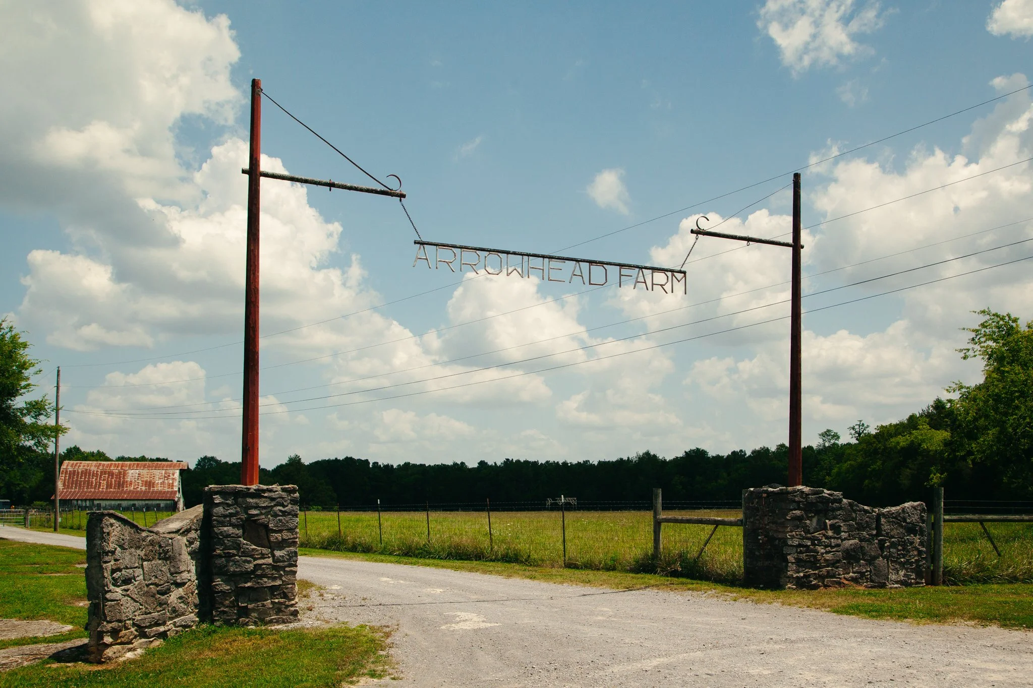 Entrance sign for Arrohead Farm with two tall red metal poles and a hanging metal sign, situated in a rural setting with a grassy field, stone pillars, and a partly cloudy sky.
