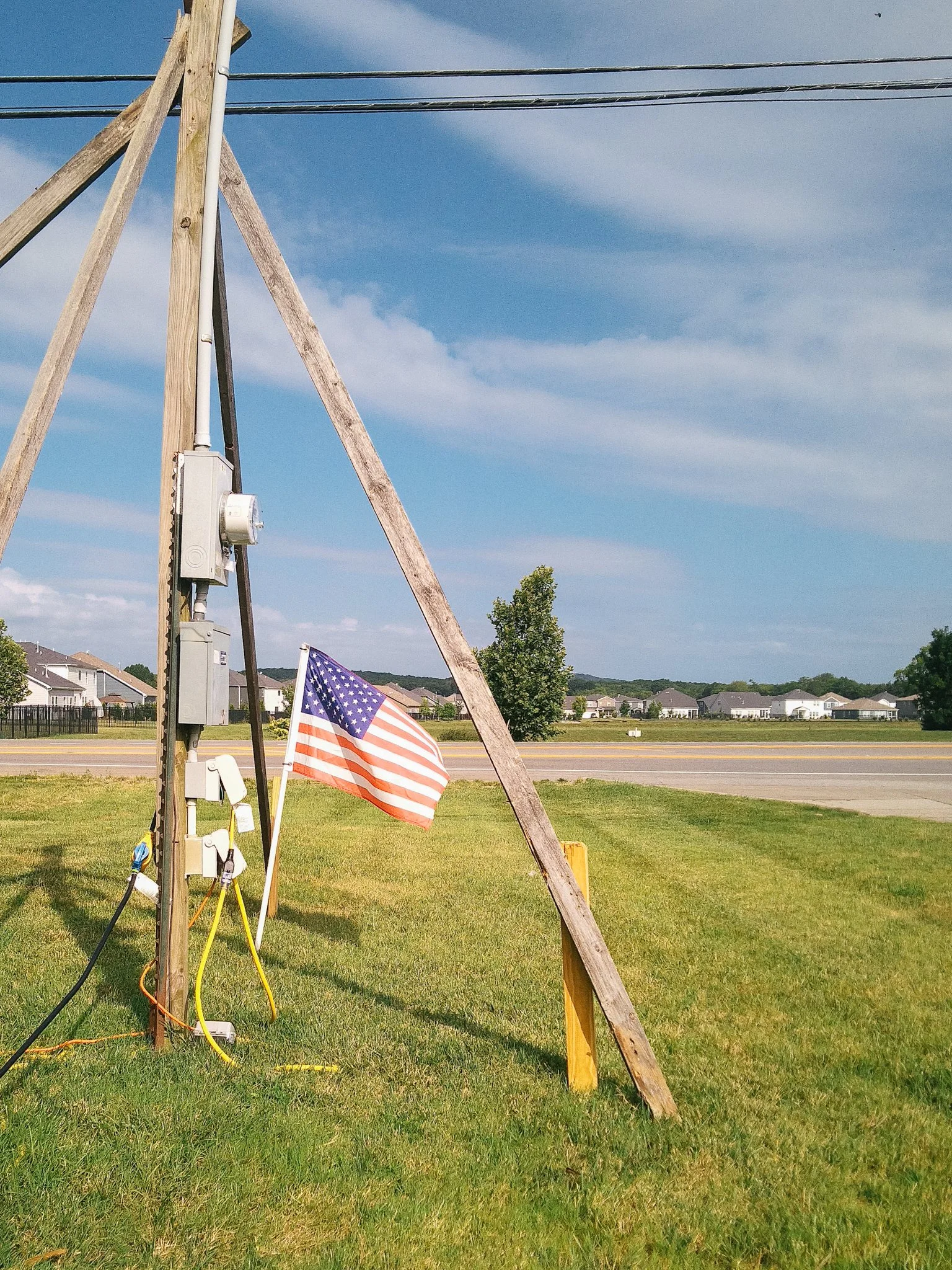 A partly set up electrical pole with wires extending across the sky, a small American flag attached to the pole, a patch of green grass, and residential houses in the background under a blue sky with clouds.