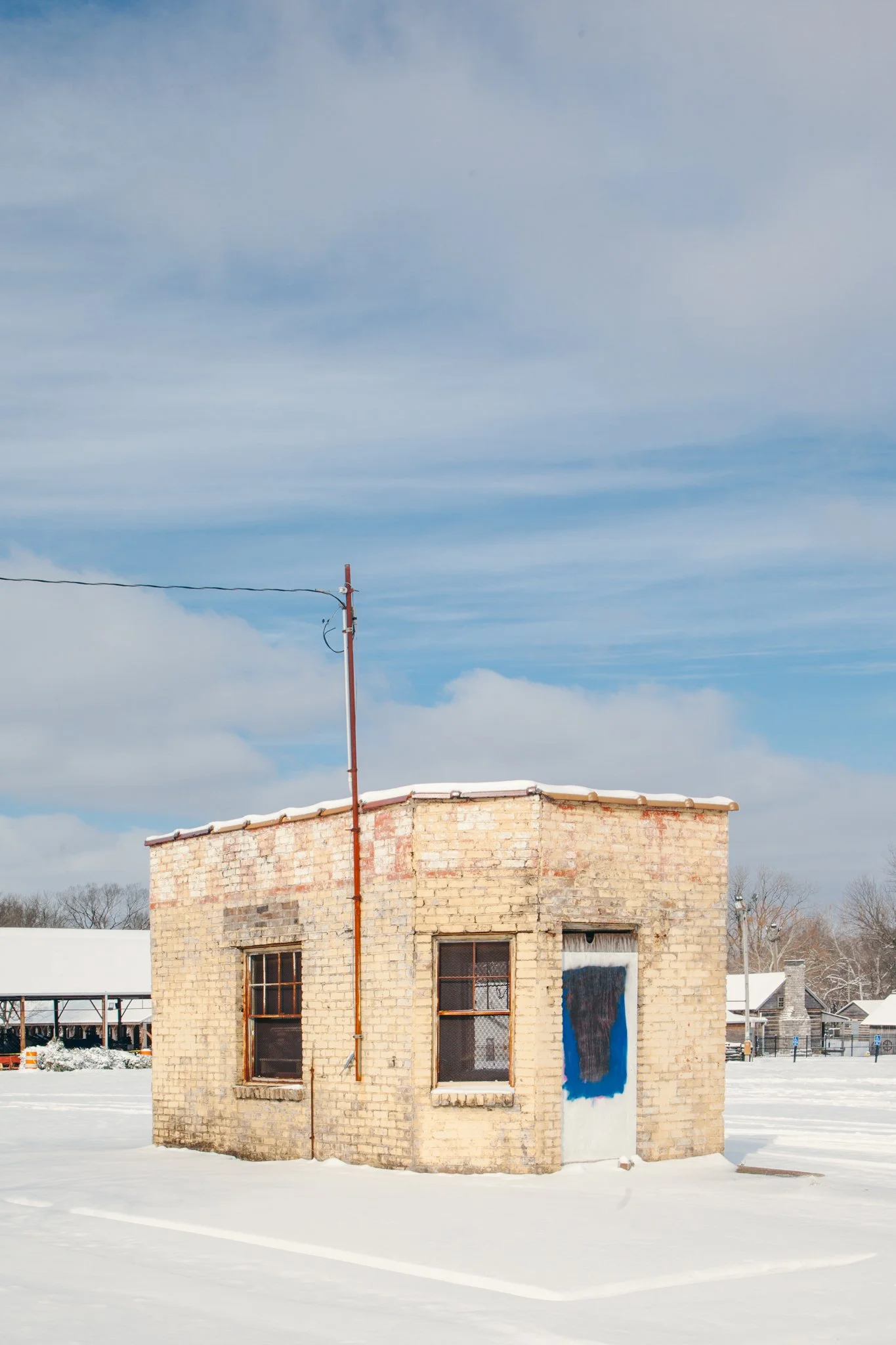 A small brick building with two windows and an open door, snow on the roof and ground, and a painted mural on the side wall, under a partly cloudy sky.