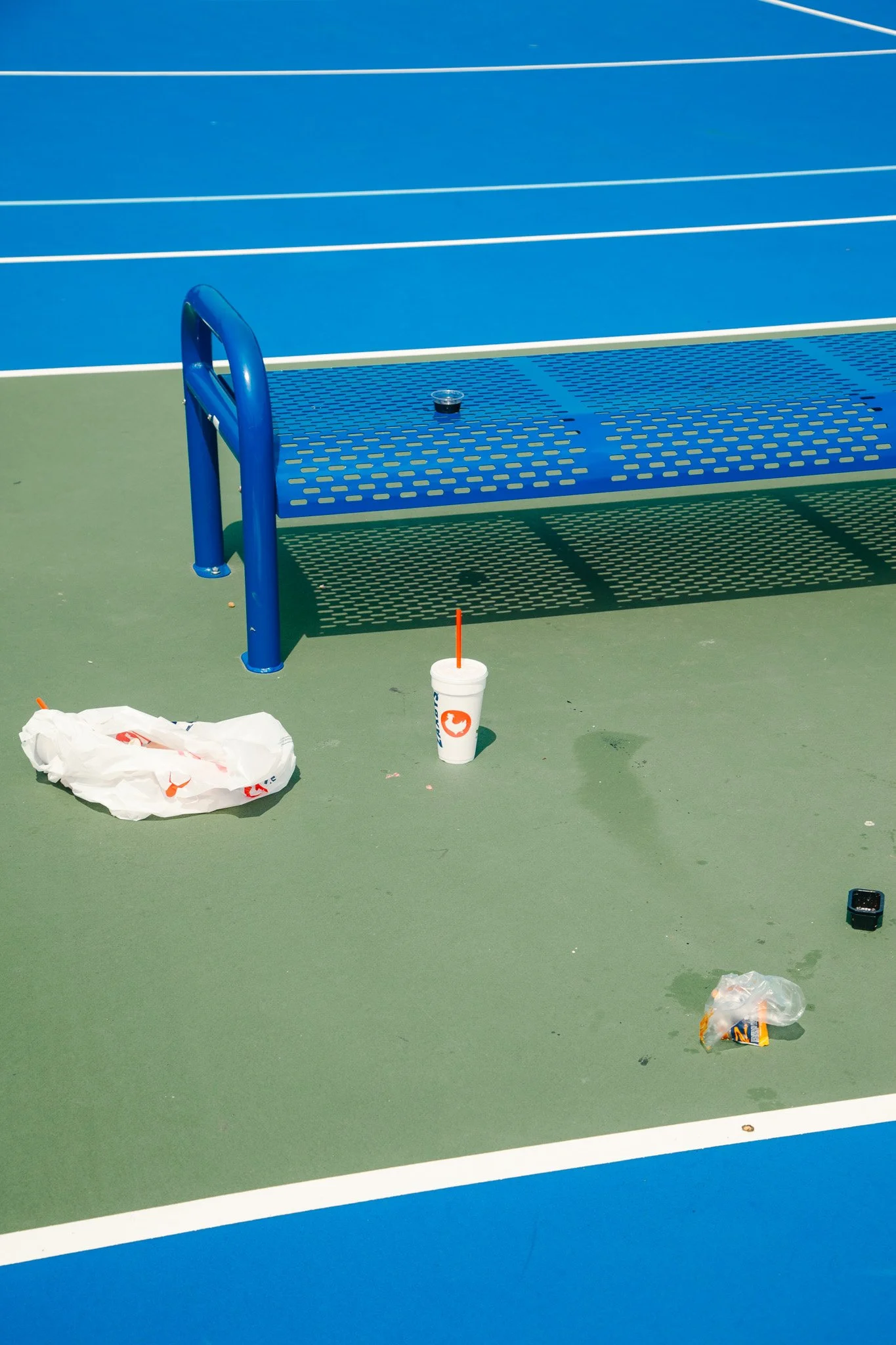 Empty tennis court with a blue bench and scattered trash including a fast food drink cup, trash bags, and a small black object on the court's surface.