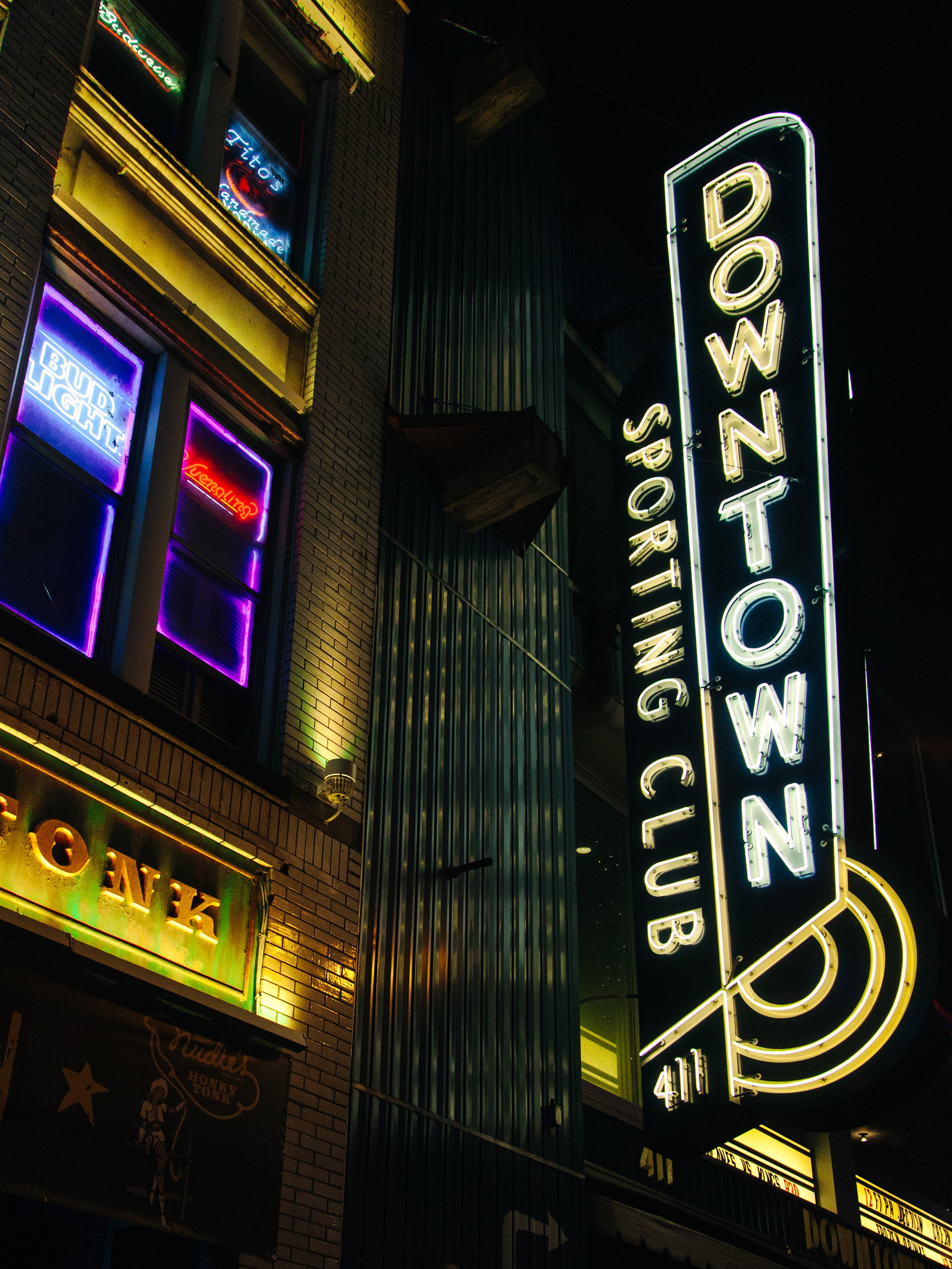Nighttime neon-lit sign for Downtown Sports Bar, with smaller neon signs for other businesses on the building, including a Bud Light sign and a sign with the word 'squid'.