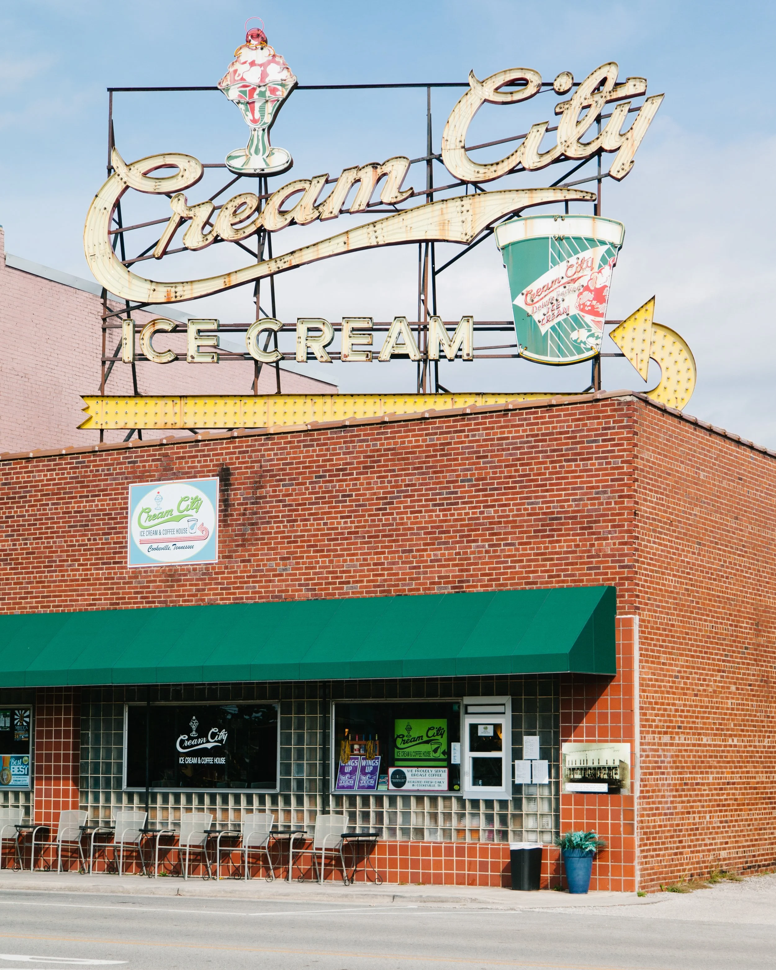 Exterior view of Cream City ice cream and coffee house with a large neon sign on top of the brick building, displaying the store name, ice cream, and a cup of ice cream. There are chairs and tables outside, and a green awning covers the entrance.