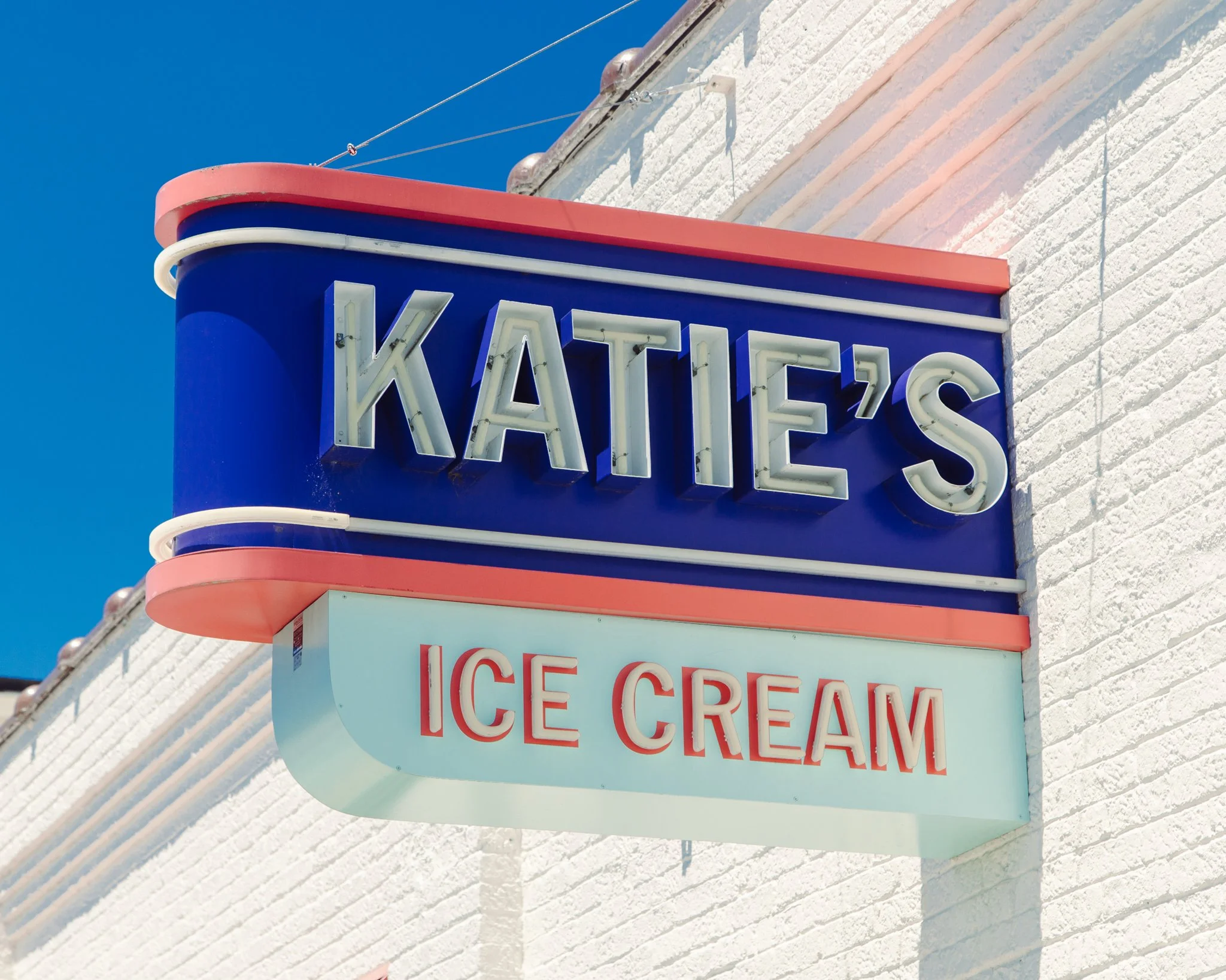 Close-up of a neon sign on a white brick building that reads 'KATIE'S ICE CREAM' with a blue background sky.