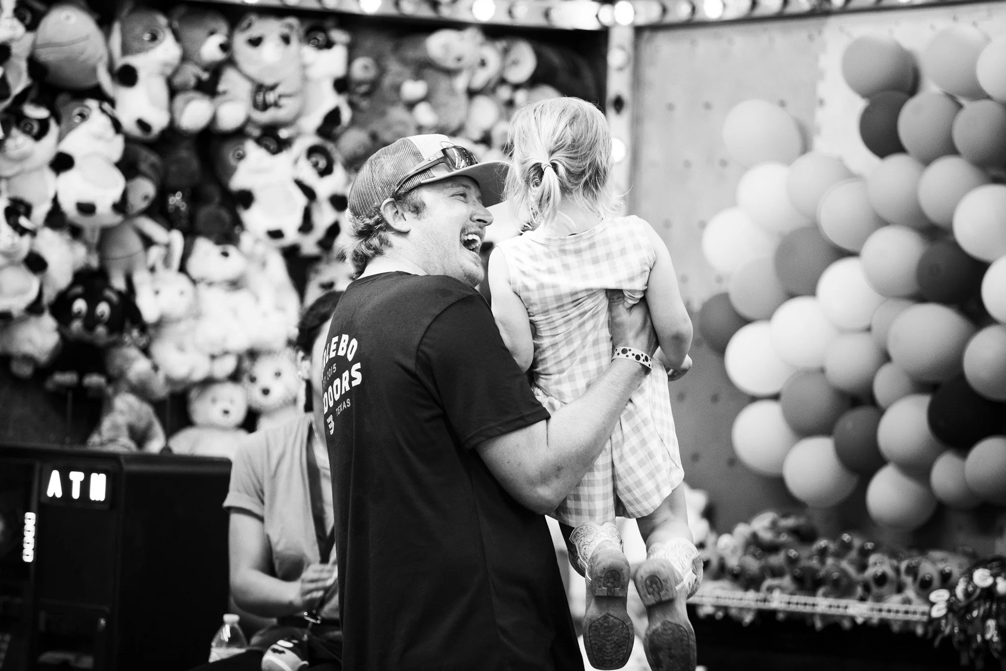 A man with a baseball cap and sunglasses on his head is smiling and holding a blonde girl in a checkered dress in a toy store. The background shows stuffed animals and balloons.