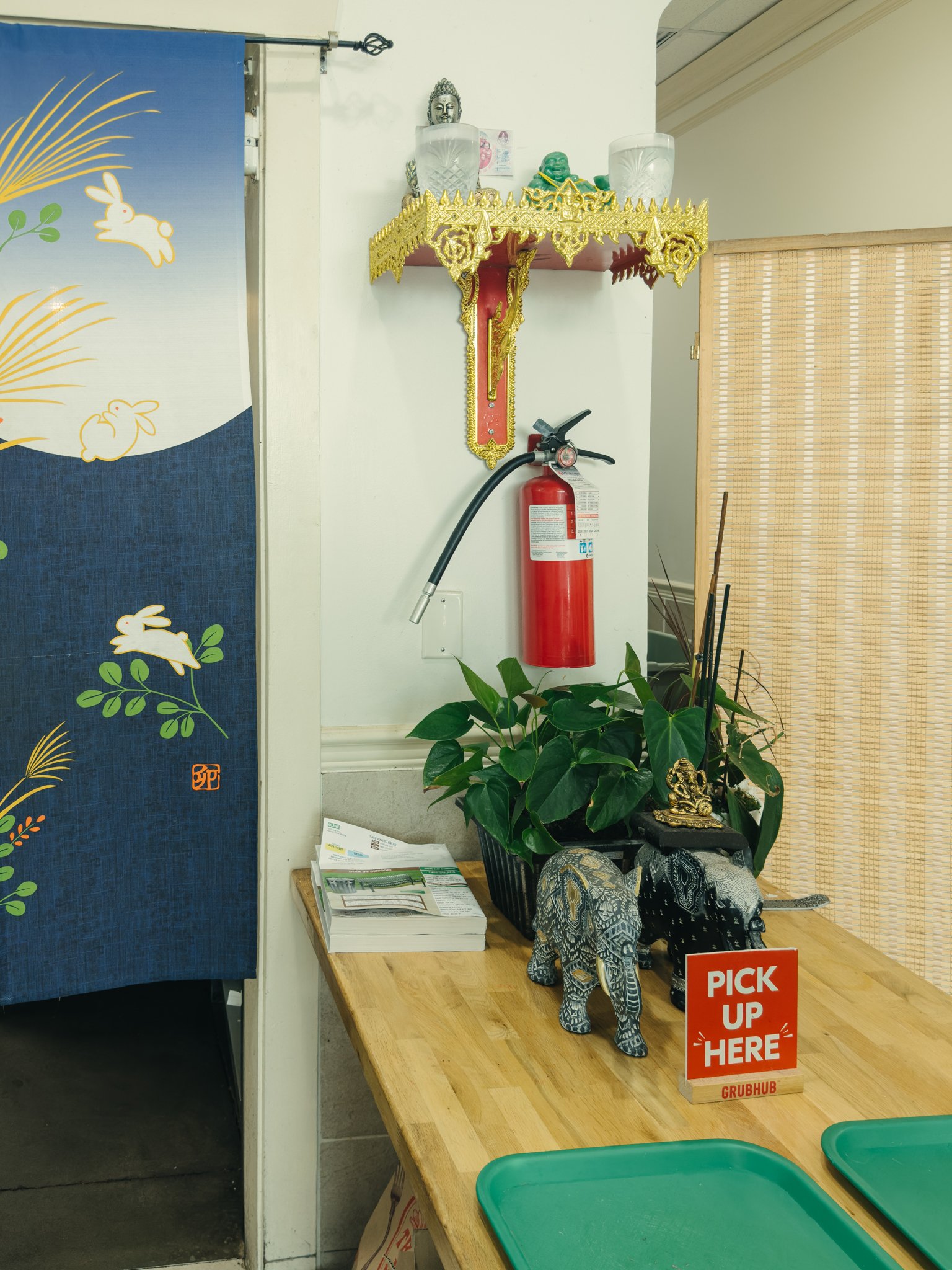A wooden table with a small decorative elephant sculpture, a plant, and a stack of papers. There is a red sign that says 'PICK UP HERE' and 'GRUBHUB'. Behind the table, a colorful prayer altar with Buddha and other statues, candles, and incense. To t