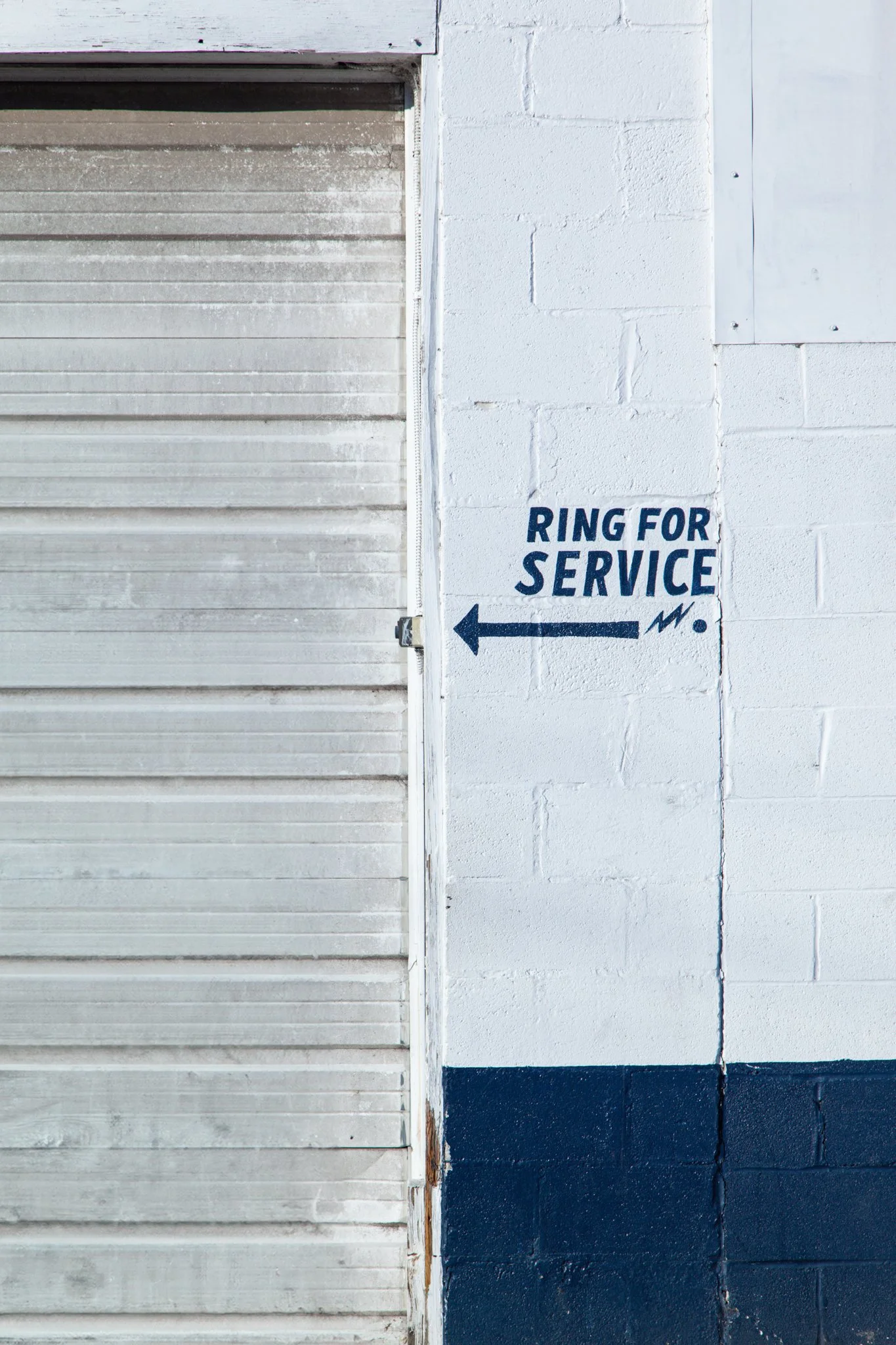 A metal garage door next to a white brick wall with a sign that reads 'Ring for Service' with an arrow pointing left, painted in blue.