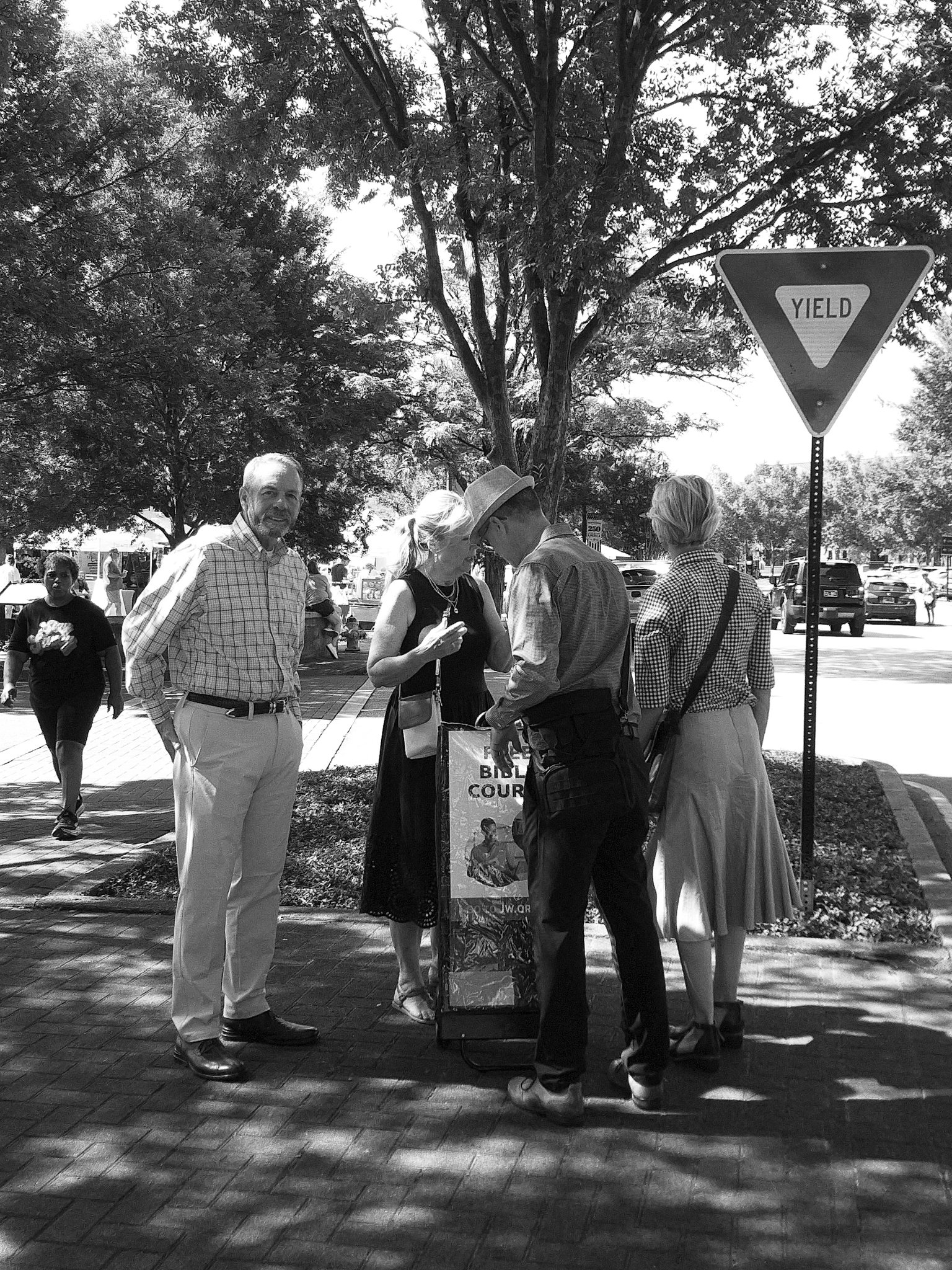 Group of people standing outdoors near a street corner with a 'Yield' traffic sign, trees, and parked cars in the background, engaged in conversation.