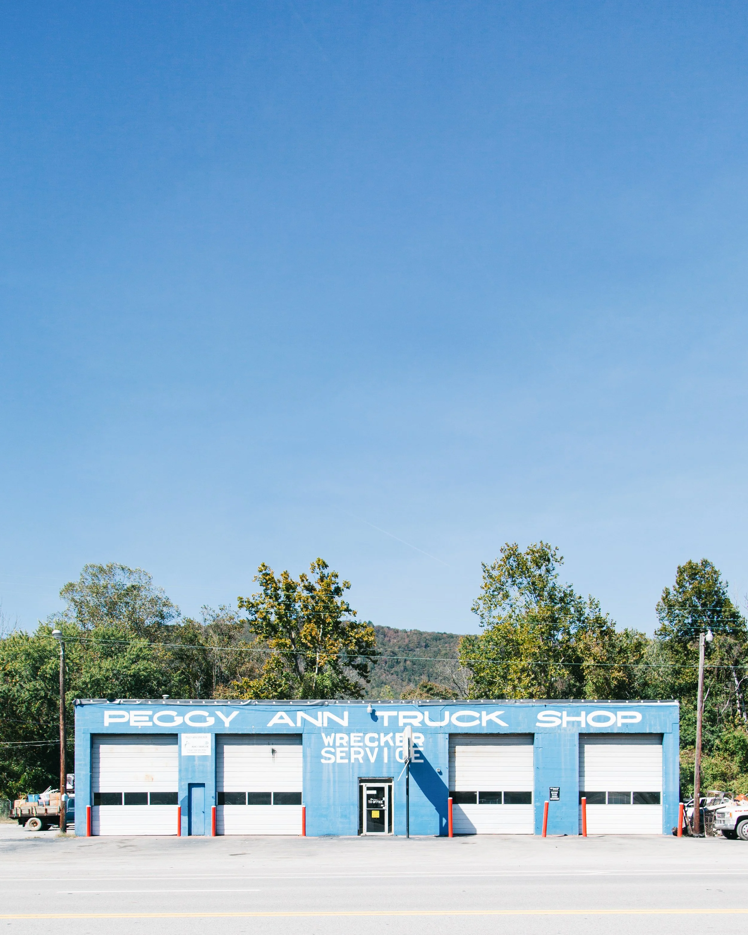 A blue building with a sign reading 'Peggy Ann Truck Shop Wrecker Service' in front of trees under a clear blue sky.