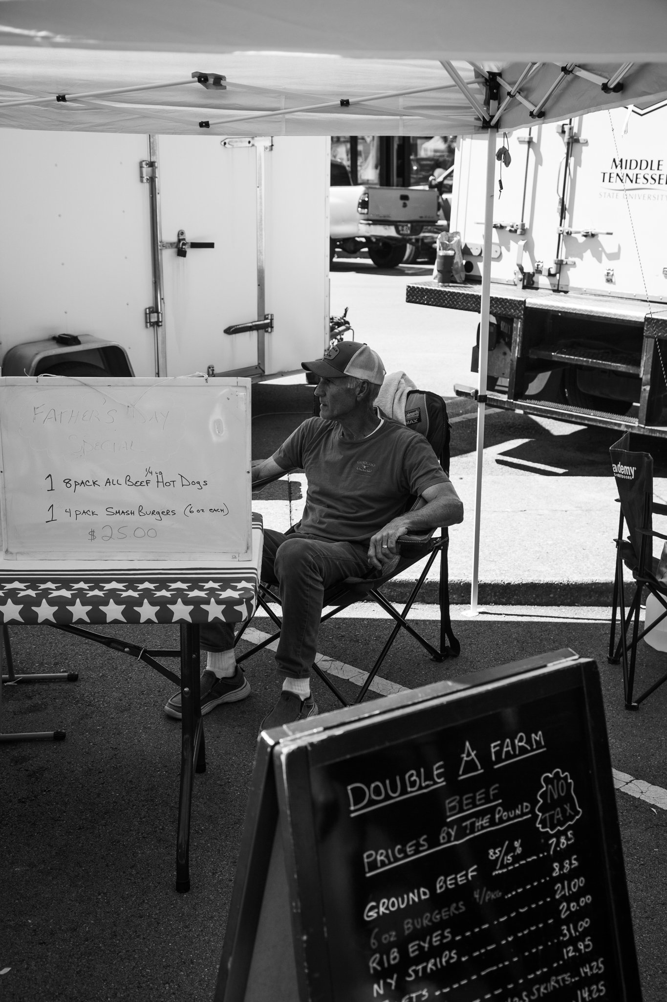 Black and white photo of a man sitting under a canopy at an outdoor food stand with a handwritten menu sign listing various types of beef and prices for a Father's Day special.