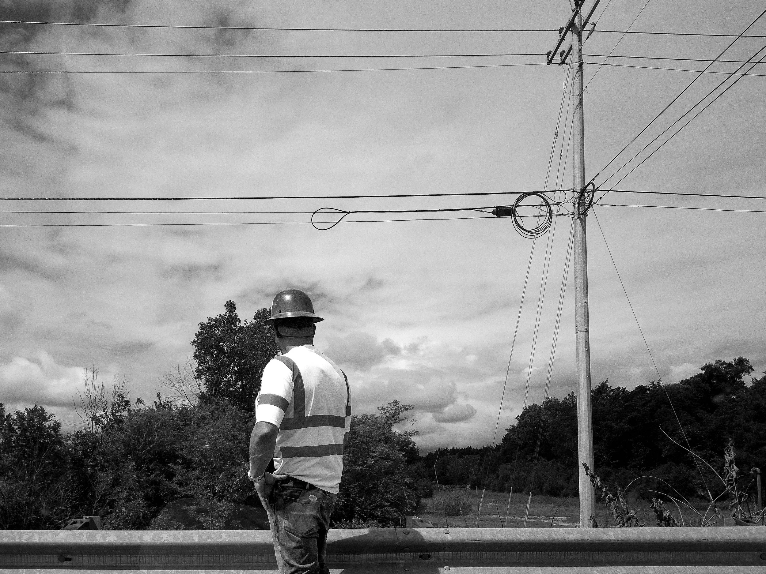 A worker wearing a hard hat and reflective vest is standing outdoors near electrical poles and wires, with trees and a cloudy sky in the background.