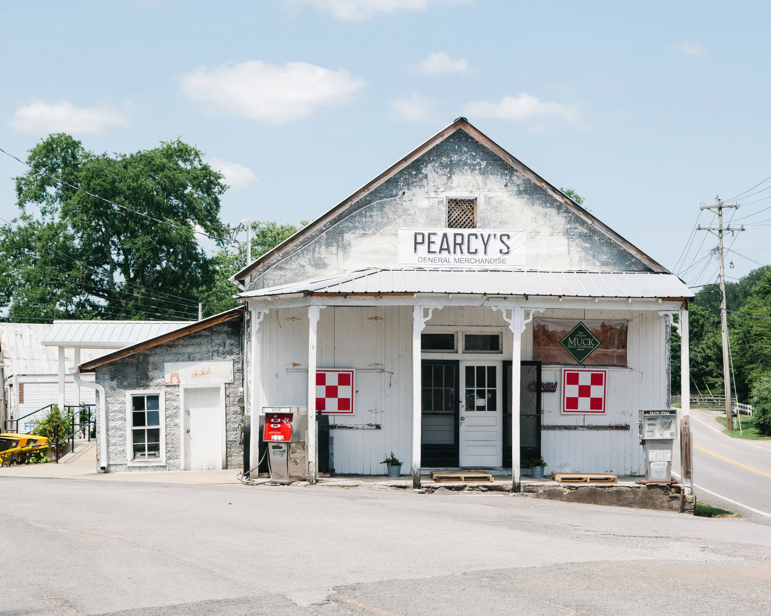 An old, weathered building with a sign that reads 'PEARCY'S GENERAL MERCHANDISE' on the front. The building has a metal roof and a porch with decorative trim. There are trees and utility poles in the background, and the sky is mostly clear with some 