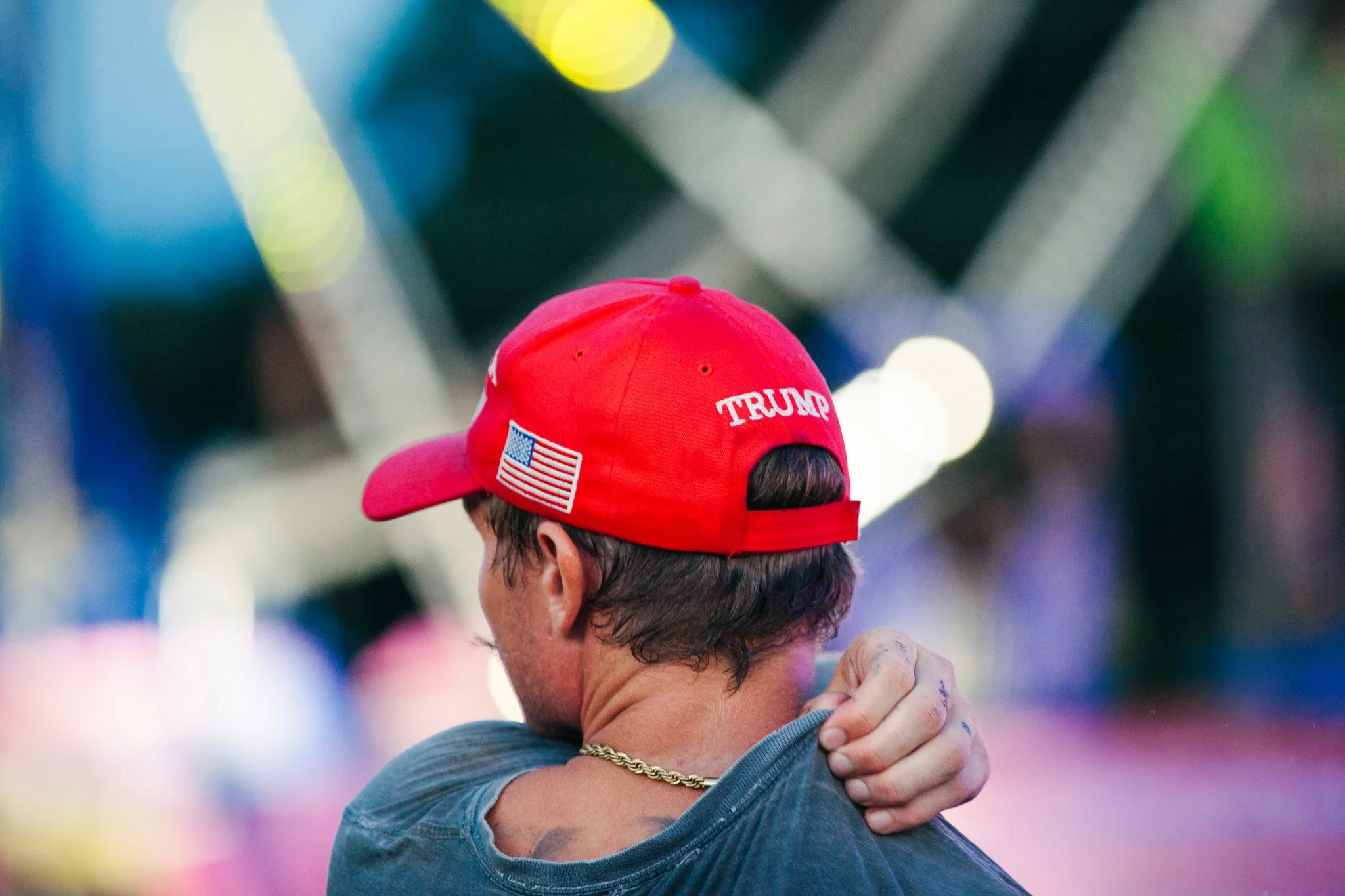 Rear view of a man wearing a red Trump cap with an American flag logo, hugging another person with one hand visible on their shoulder, in a setting with colorful blurred lights.
