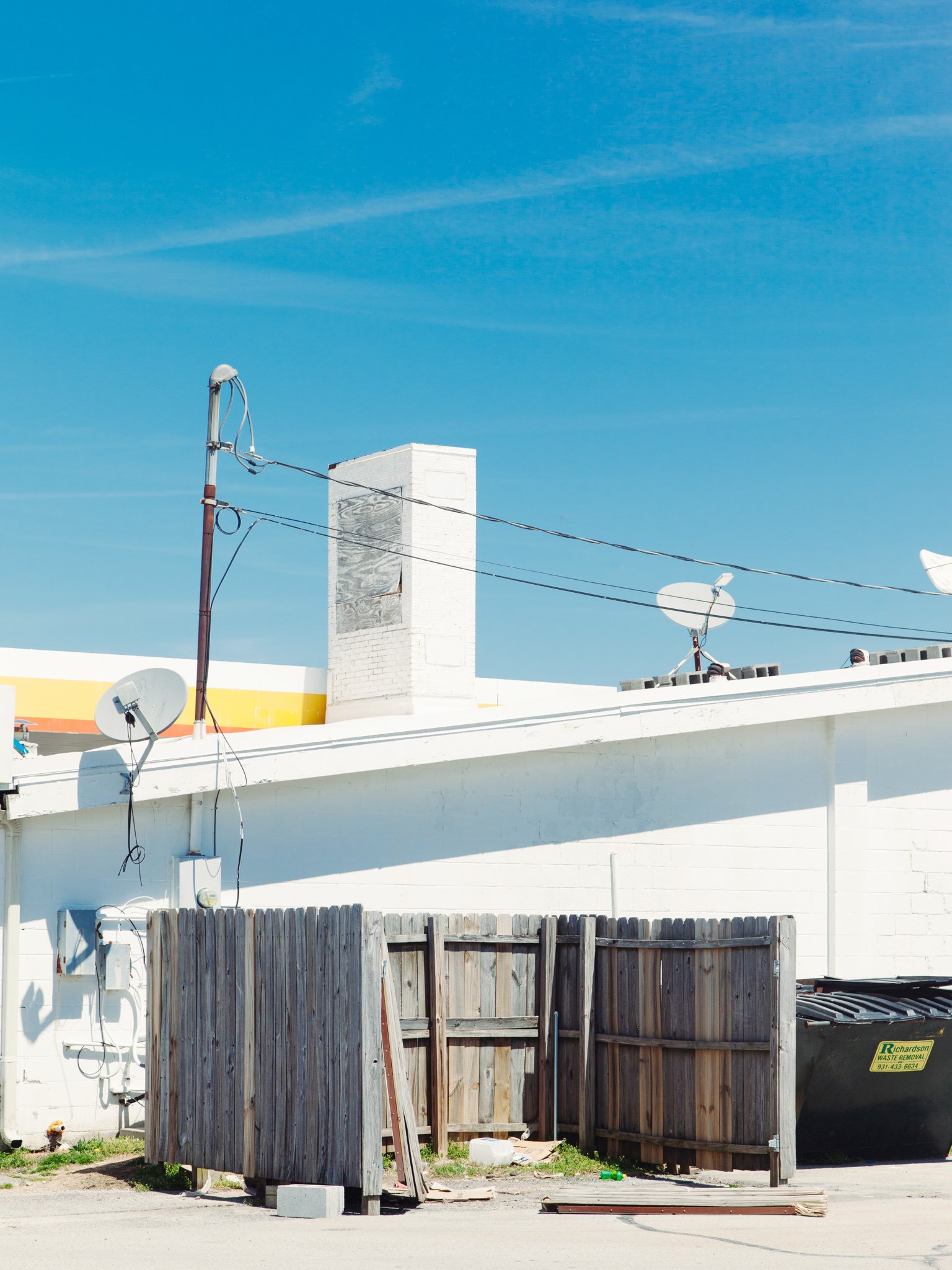 View of a rooftop with a chimney, satellite dishes, and electrical wires against a blue sky. In the foreground, there is a wooden fence with a gate and some construction debris.