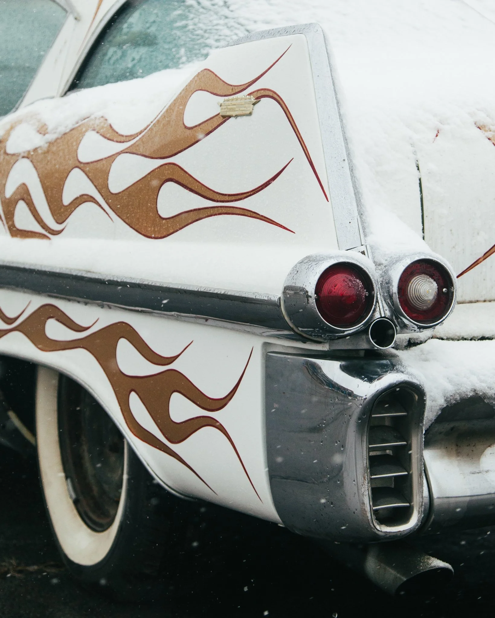Close-up of the rear end of a white luxury sports car, showing taillights, rear bumper, and part of the trunk with flames painted on the side, parked in a snowy environment.
