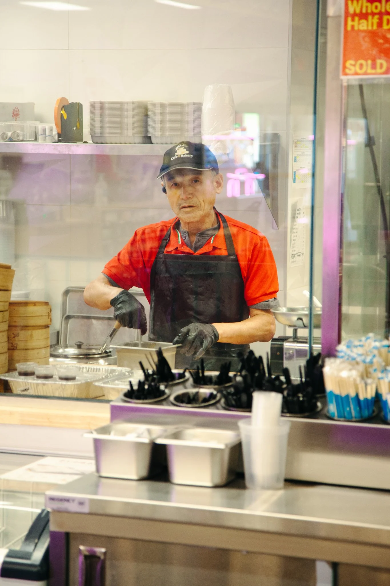 A man wearing a black cap, red shirt, and apron preparing food behind a counter in a restaurant kitchen, seen through a glass window at Pan-Asia Supermarket in Antioch, TN