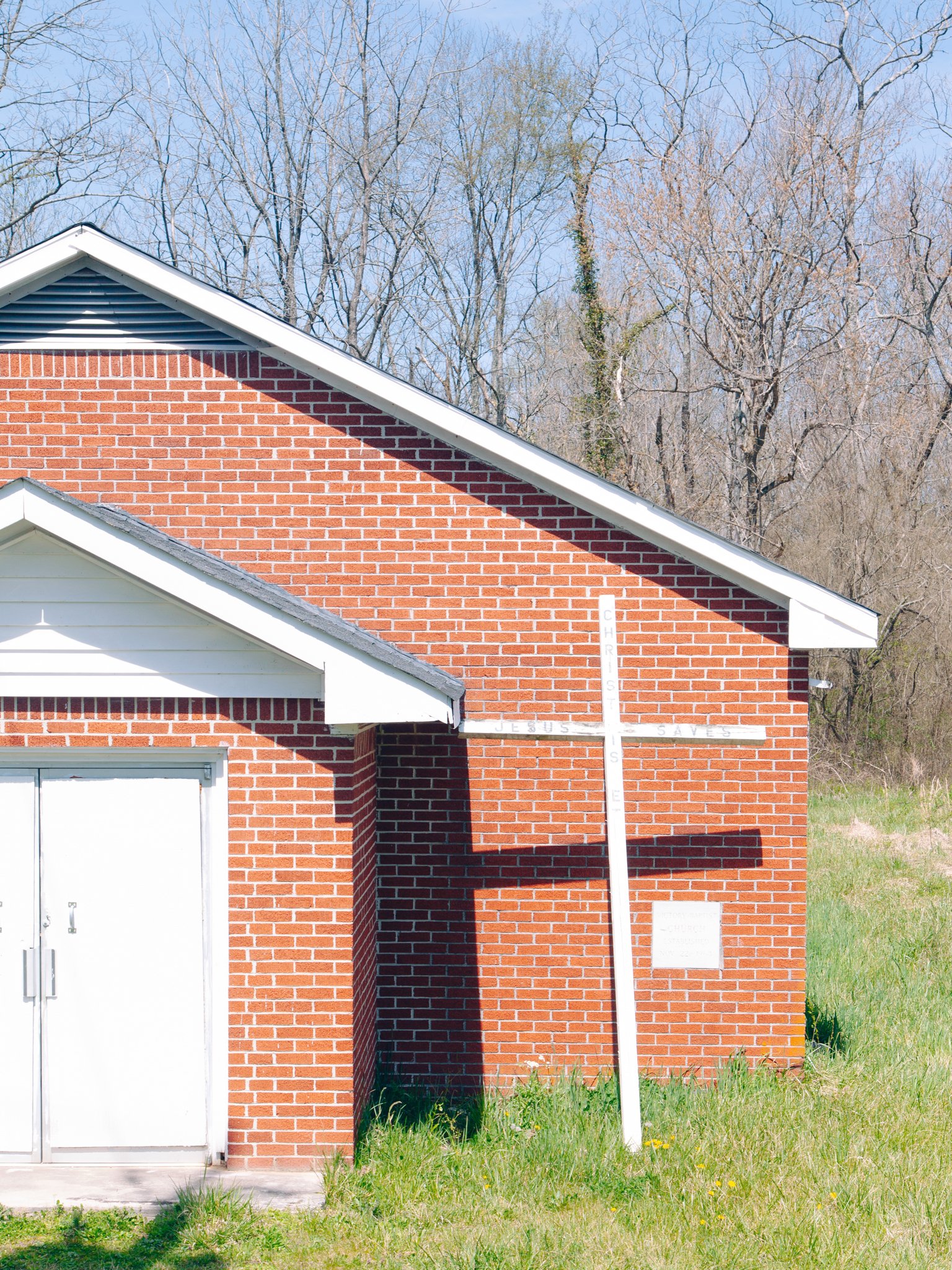 A brick building with a white garage door and white trim under a clear blue sky. A white cross with words is in front of the building, and there is grassy land with small yellow flowers at the base.