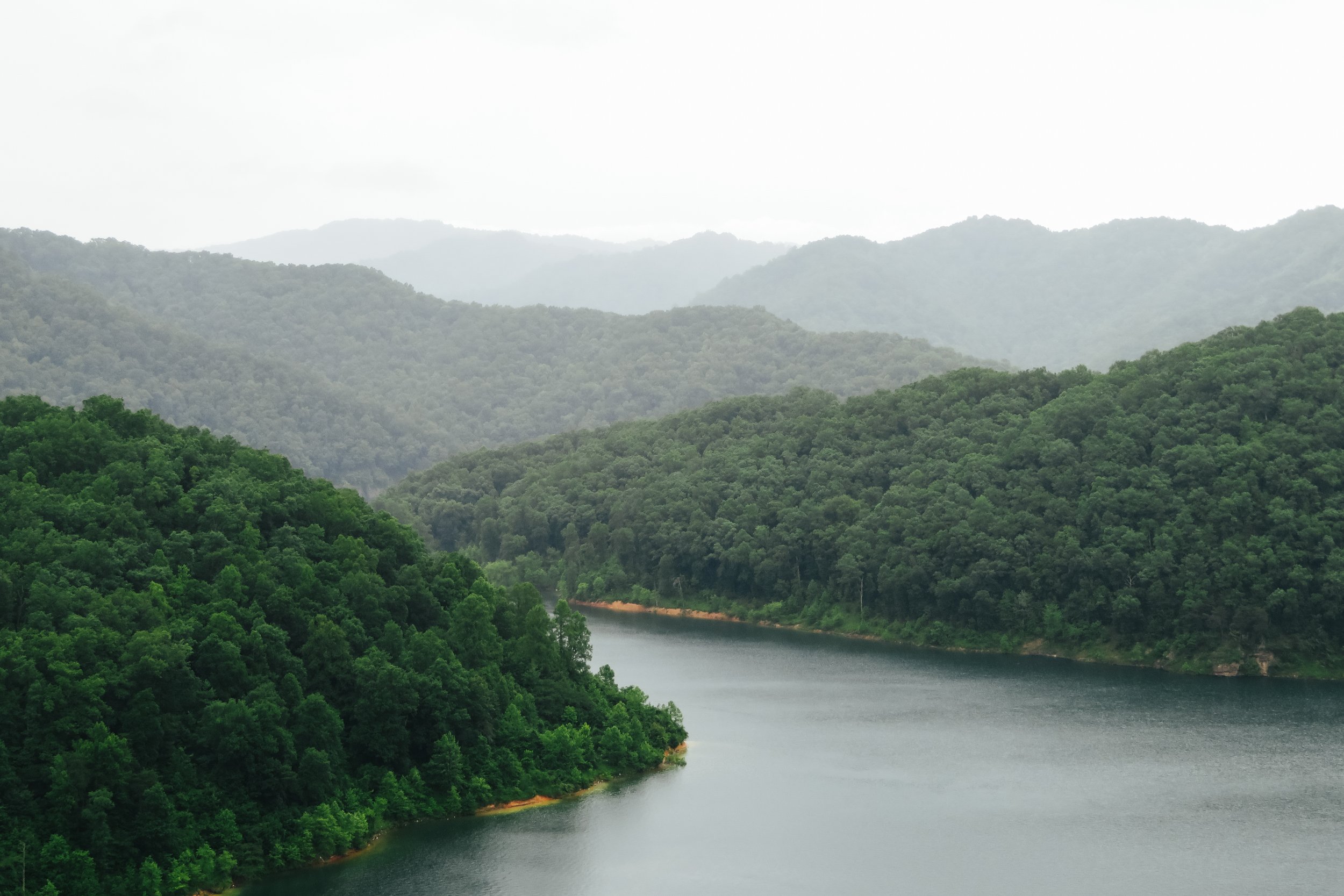 Lush green forested hills surrounding a calm lake with misty mountains in the background.