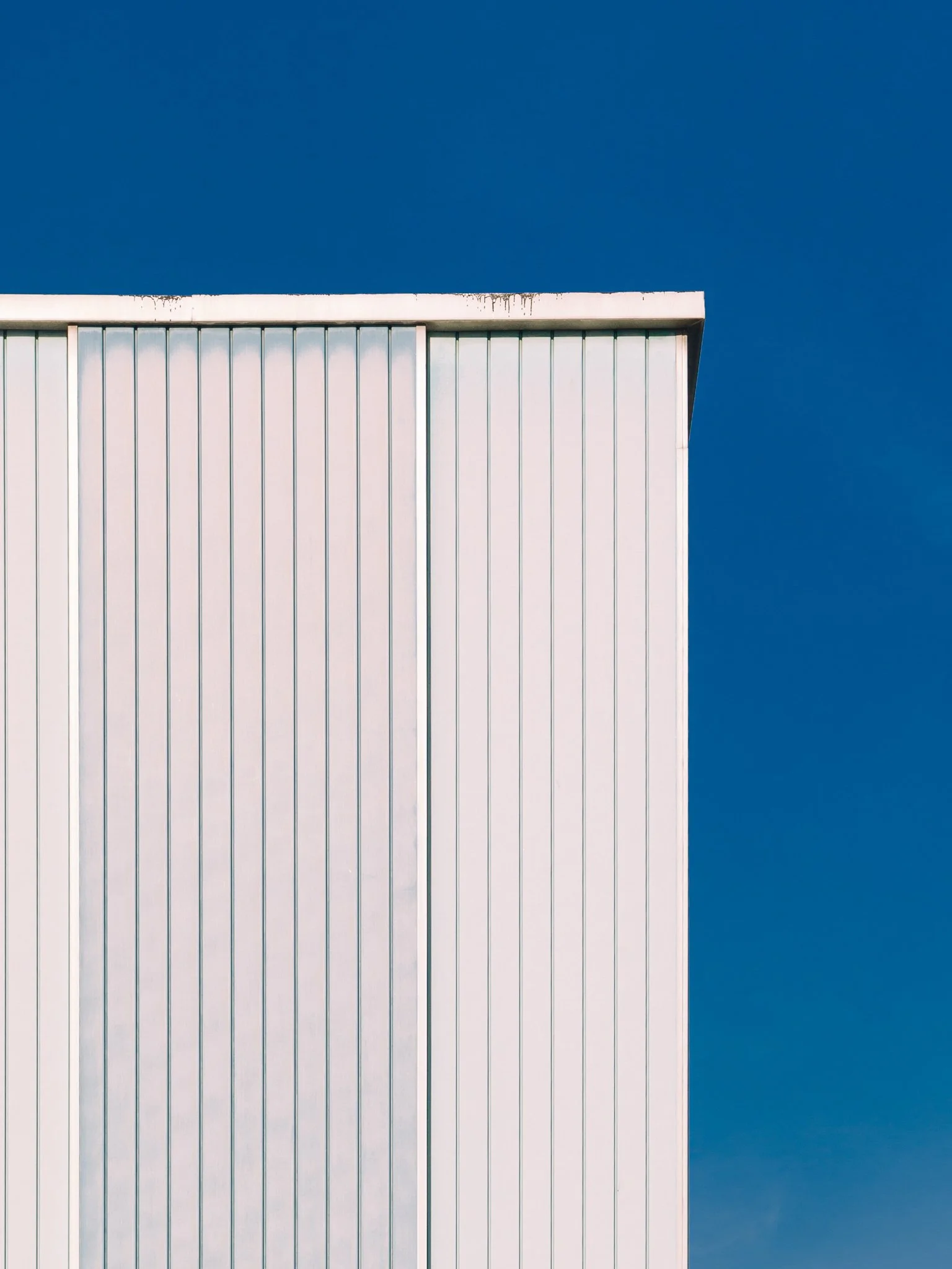A close-up of a modern building's white metal panel facade with vertical ridges, against a clear deep blue sky.