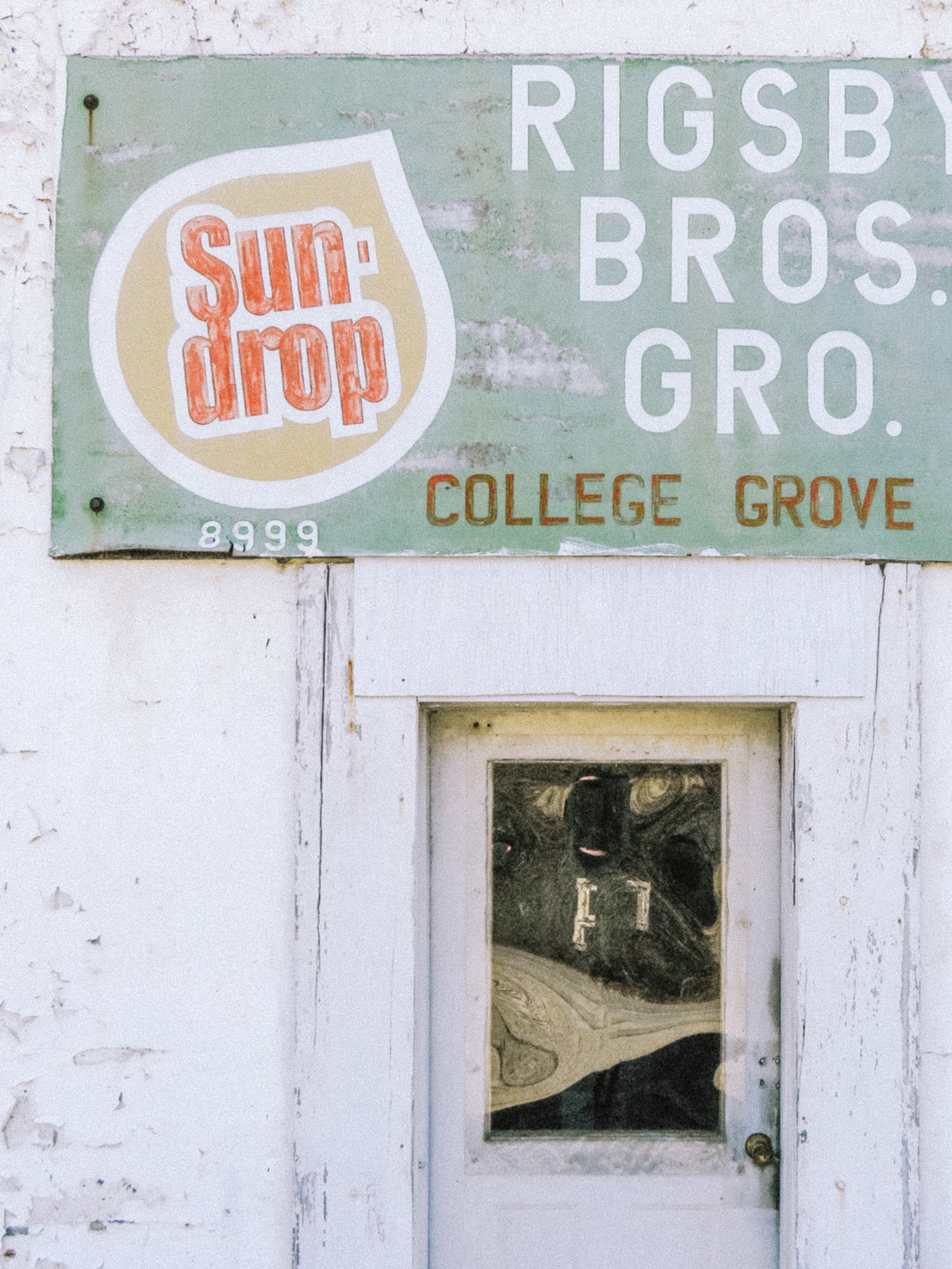 A green sign with the Sun Drop logo and the words 'Rigsby Bros. Gro.' and 'College Grove.' There is a small window below the sign, showing a reflection of the person taking the photo and an interior with a marble-like countertop.