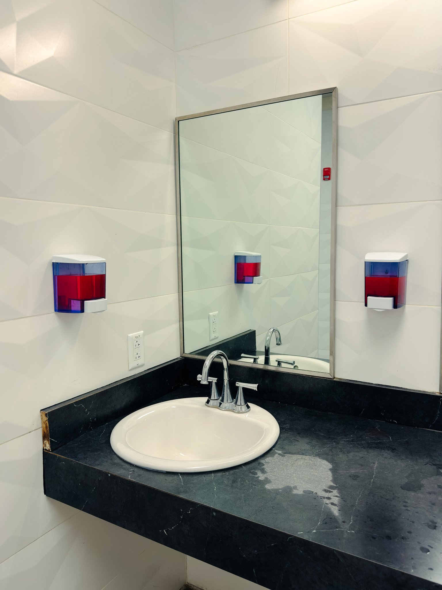 Bathroom bathroom with a mirror above a black marble countertop, a white sink, and two wall-mounted soap dispensers with red and blue caps.