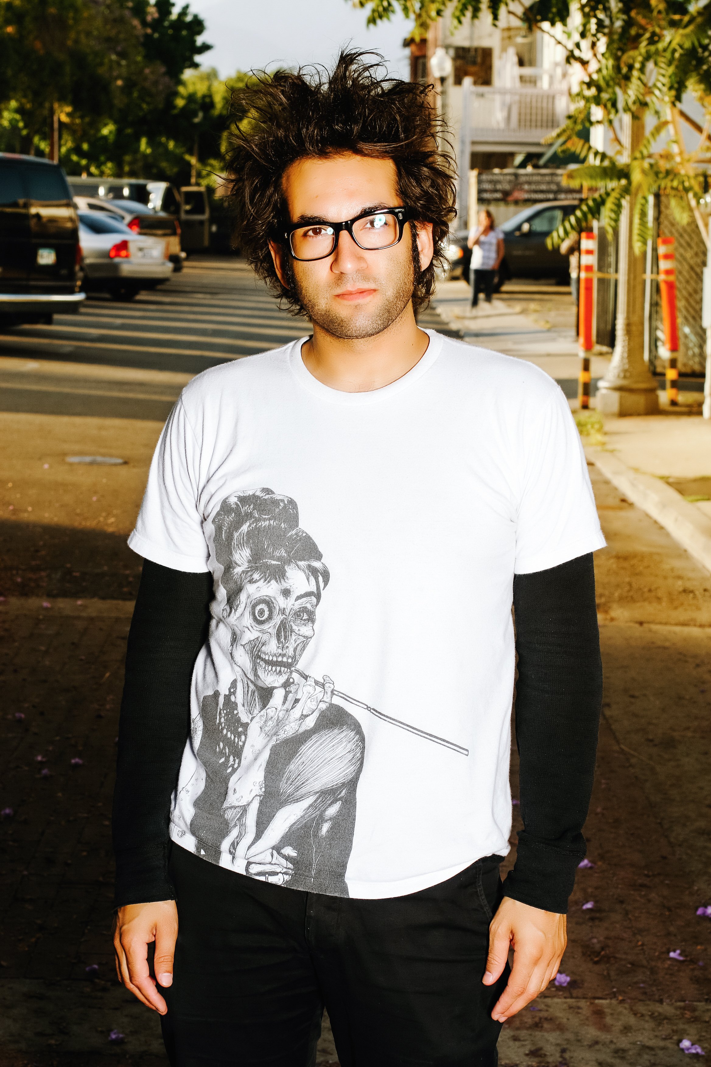 A man with curly dark hair, glasses, and a white t-shirt with a skeleton woman playing a flute stands on a city sidewalk with parked cars, trees, and a woman in the background.