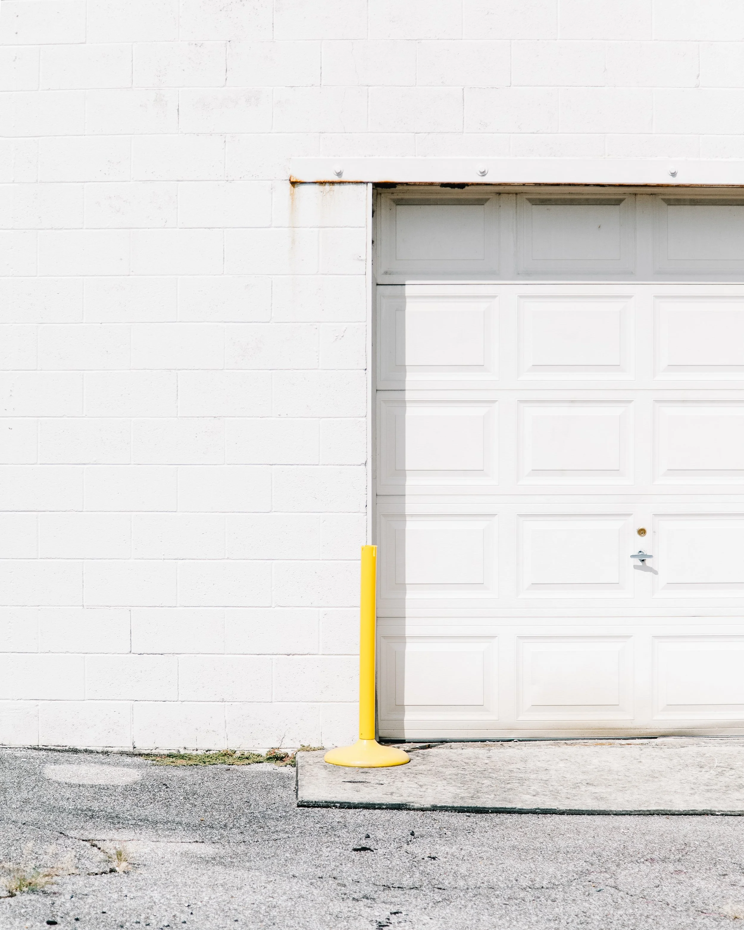 A white garage door set within a white brick wall, with a yellow safety pole in front of the door.