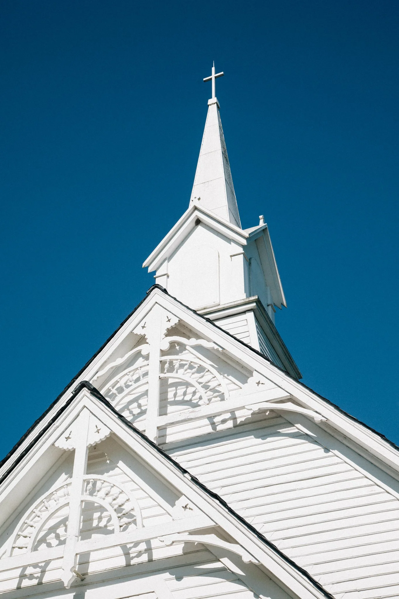 White church steeple with a cross on top against a blue sky, featuring decorative architectural details.