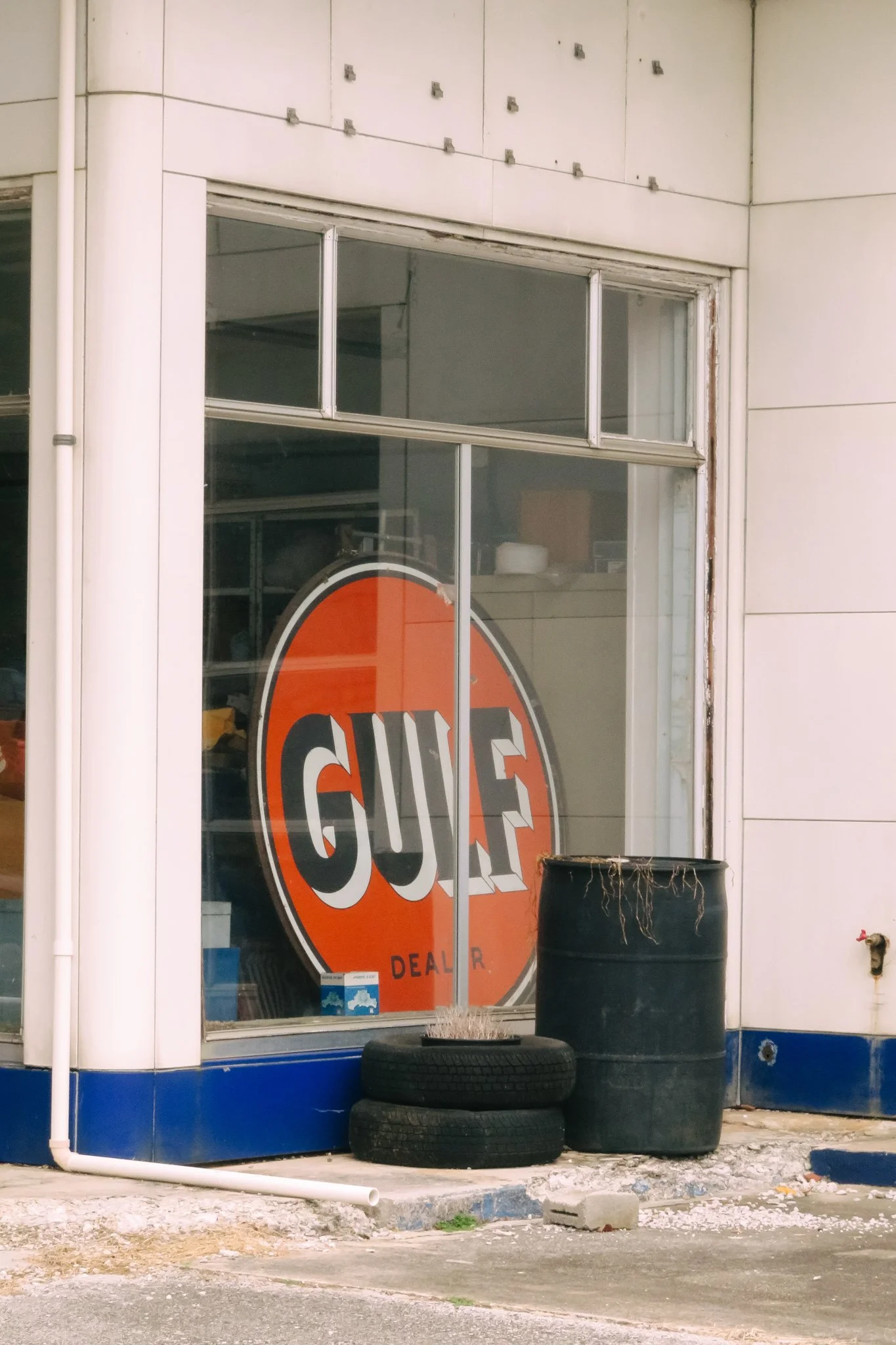 View of a storefront with a large, orange and black circular sign that reads 'GNA' and 'DEAL R.' in black text, surrounded by stacked tires, a black trash barrel, and a lower section of blue wall with broken concrete and debris outside.