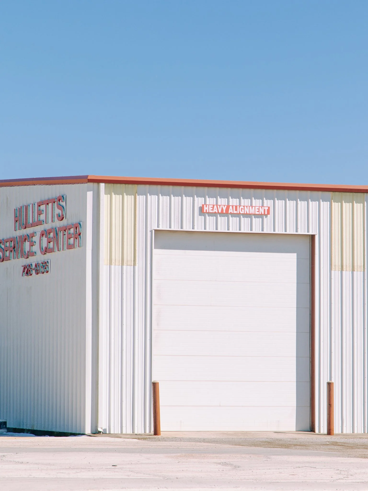 A white industrial building with a garage door, orange roof edging, and a sign that says 'Heavy Alignment' above the garage door. There are two orange bollards in front of the building and a sidewalk, with a clear blue sky above.