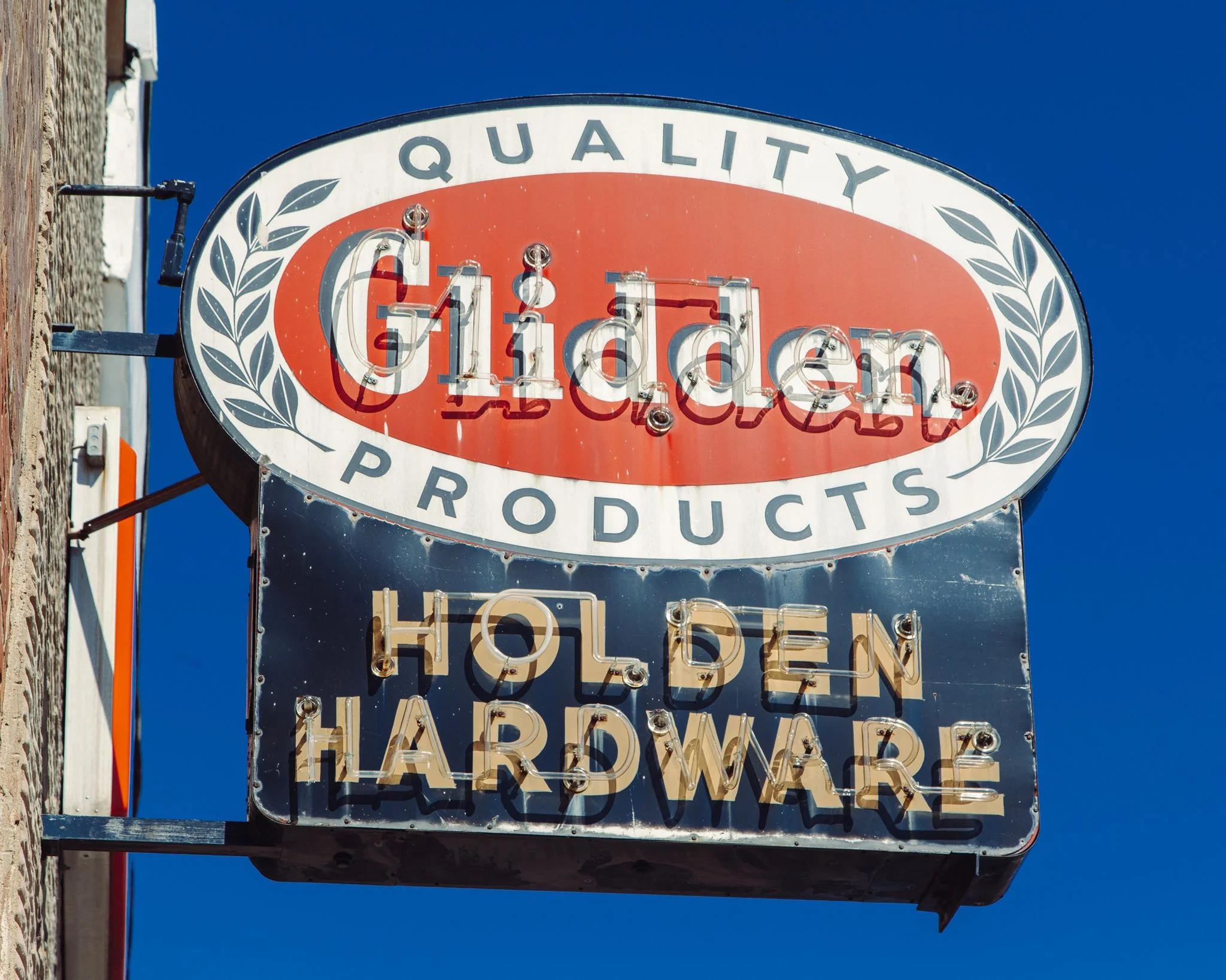 A vintage-style sign reading 'Quality C11den Products' with neon letters, mounted on a building against a bright blue sky.