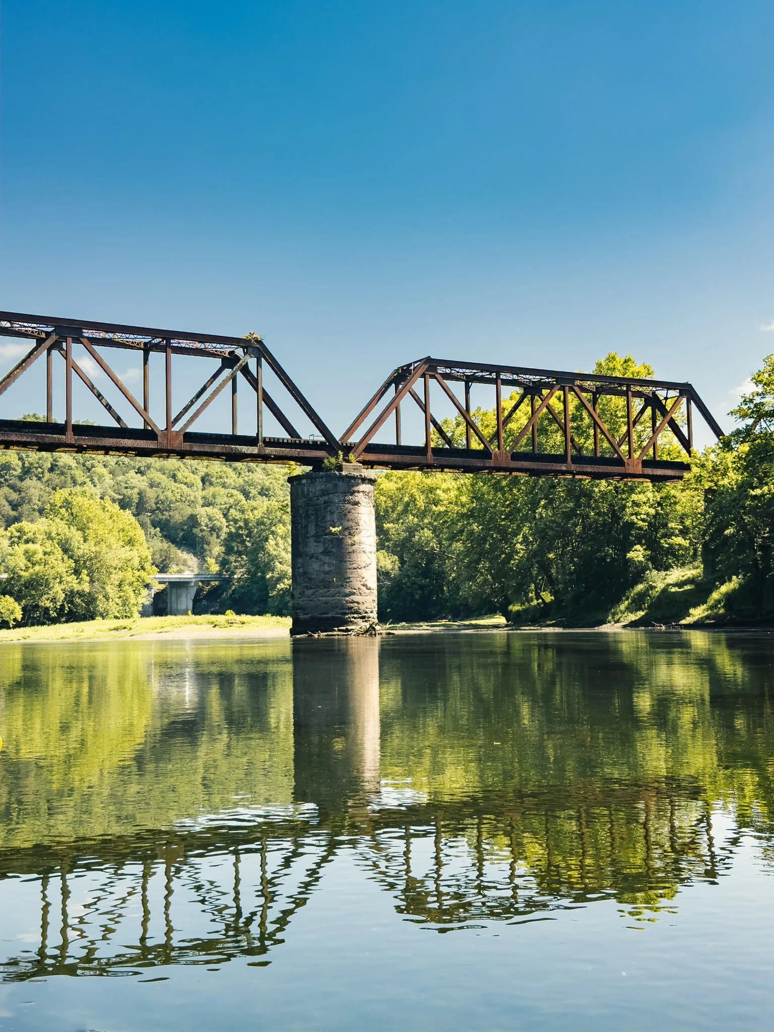 An old railroad bridge crossing over a calm river, surrounded by lush green trees and clear blue sky.